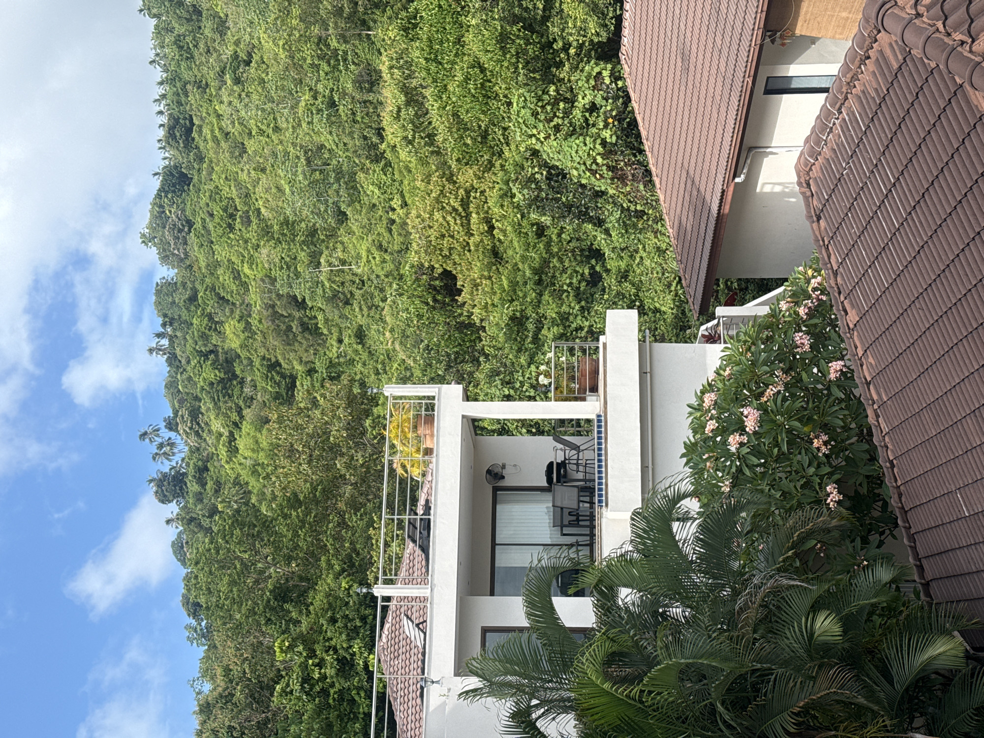 In the city of Ko Samui District, Thailand, a multi-story white building with a modern design is situated against a backdrop of lush green foliage and a clear blue sky with scattered clouds. The building features a rooftop terrace with a metal railing and potted plants, as well as a second-floor balcony with outdoor furniture, a barbecue grill, and a small pool. In the foreground, two tiled roofs of residential structures are visible. The surrounding environment is characterized by dense tropical vegetation, including palm trees and flowering bushes. The scene appears peaceful and tranquil.