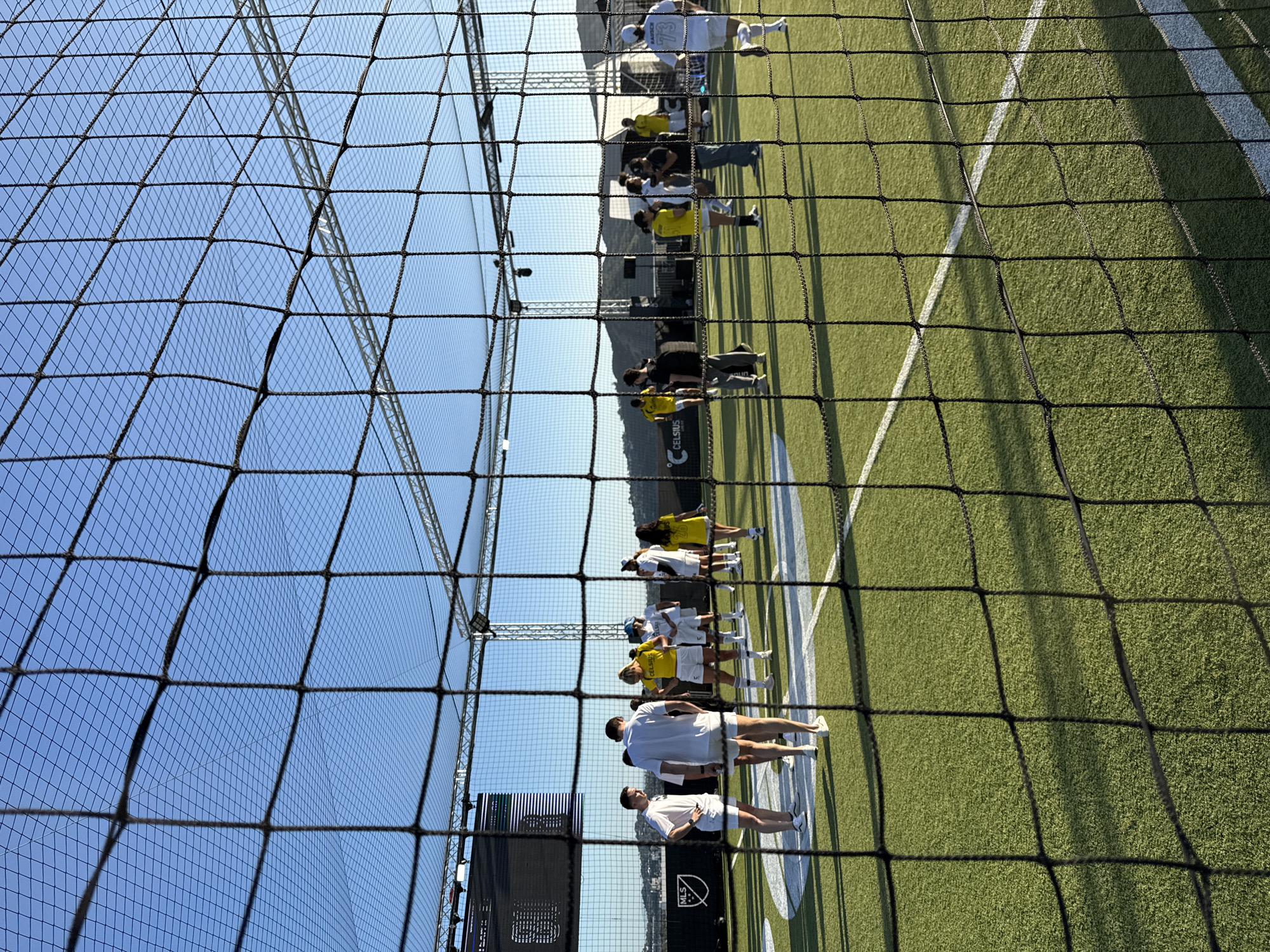 The image shows a view through a black netting onto a green field marked with white lines, likely an athletic field. People, mostly wearing white or yellow shirts, are standing on the field. In the background, there is a mountain range and what appears to be a digital screen with the MLS logo. The image is likely taken in Glendale, USA, as indicated by the MLS logo and the visible landscape.