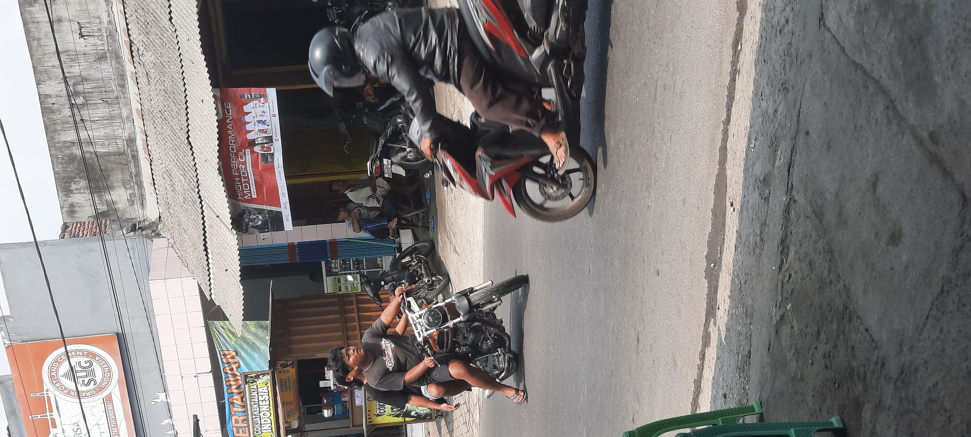 In Selopuro, Indonesia, on a sunny day, two young men are riding a motorcycle down a paved road. The driver, a young man with dark hair and wearing a grey t-shirt and shorts, is focused on the road ahead. Behind him, another young man, also with dark hair and wearing a black t-shirt and shorts, is looking towards the right. They are traveling from left to right in the frame.
In the background, a man wearing a black leather jacket and helmet is riding a red and black motorcycle from right to left. He is partially visible as he passes. Further back, several other motorcycles are parked. There are also a few men sitting on a bench near some buildings.
The buildings in the background feature signs advertising "SUG PORTLAND CEMENT INDONESIA" and "HIGH PERFORMANCE MOTOR OIL." The overall scene depicts a typical street in an Indonesian town, with commerce and transportation coexisting. The road is made of asphalt and appears to be in moderate condition, with some cracks and patches visible. The environment suggests a warm, possibly humid climate.