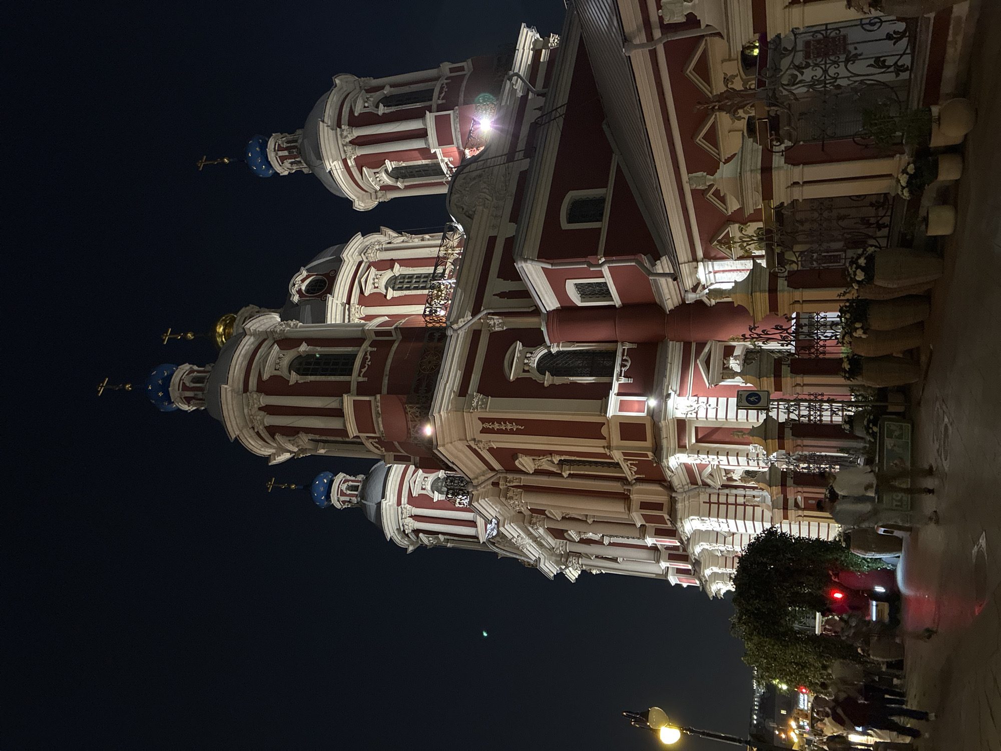 The image depicts the Saint Nicholas Naval Cathedral in Moscow, Russia at night. The cathedral features red and white architectural elements, with blue and gold domes topped with crosses. A decorative iron fence runs along the right side of the frame, adorned with potted plants and flowers. The foreground shows a dimly lit street scene with pedestrians and a vehicle with red taillights. A street lamp is visible on the bottom left. The sky above the cathedral is dark.