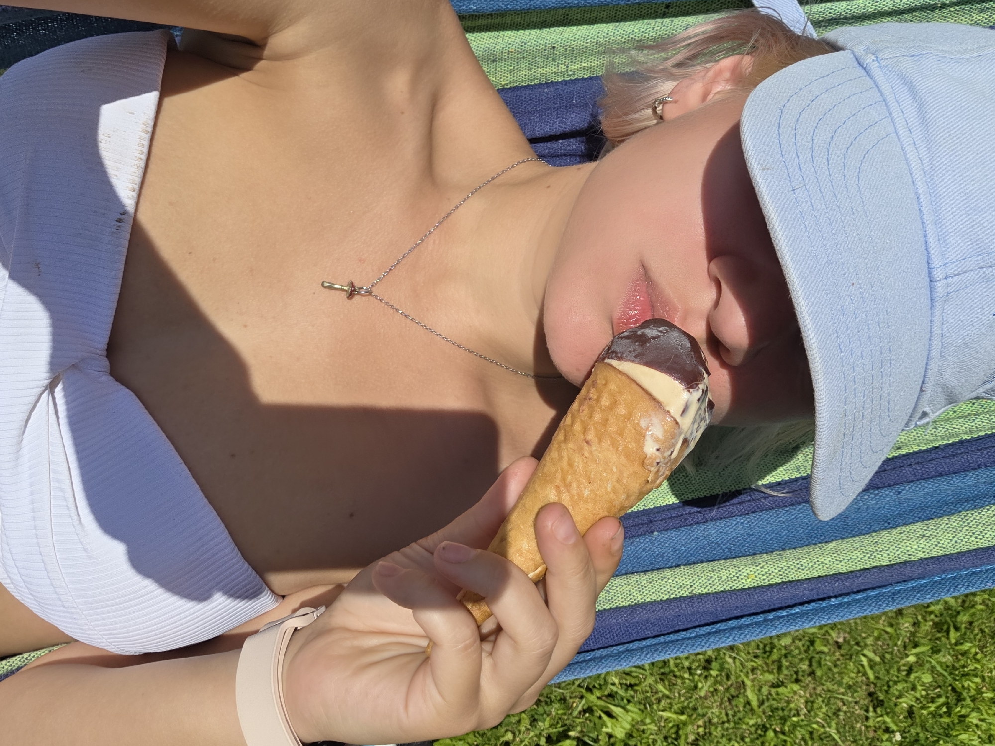 A young woman with blonde hair and a light blue baseball cap is lying in a hammock outdoors, holding an ice cream cone to her lips. She is wearing a white strapless top and a silver necklace with a mushroom pendant. The hammock is striped in blue and green, and is positioned on a lawn. The sun is shining, casting shadows on her body.