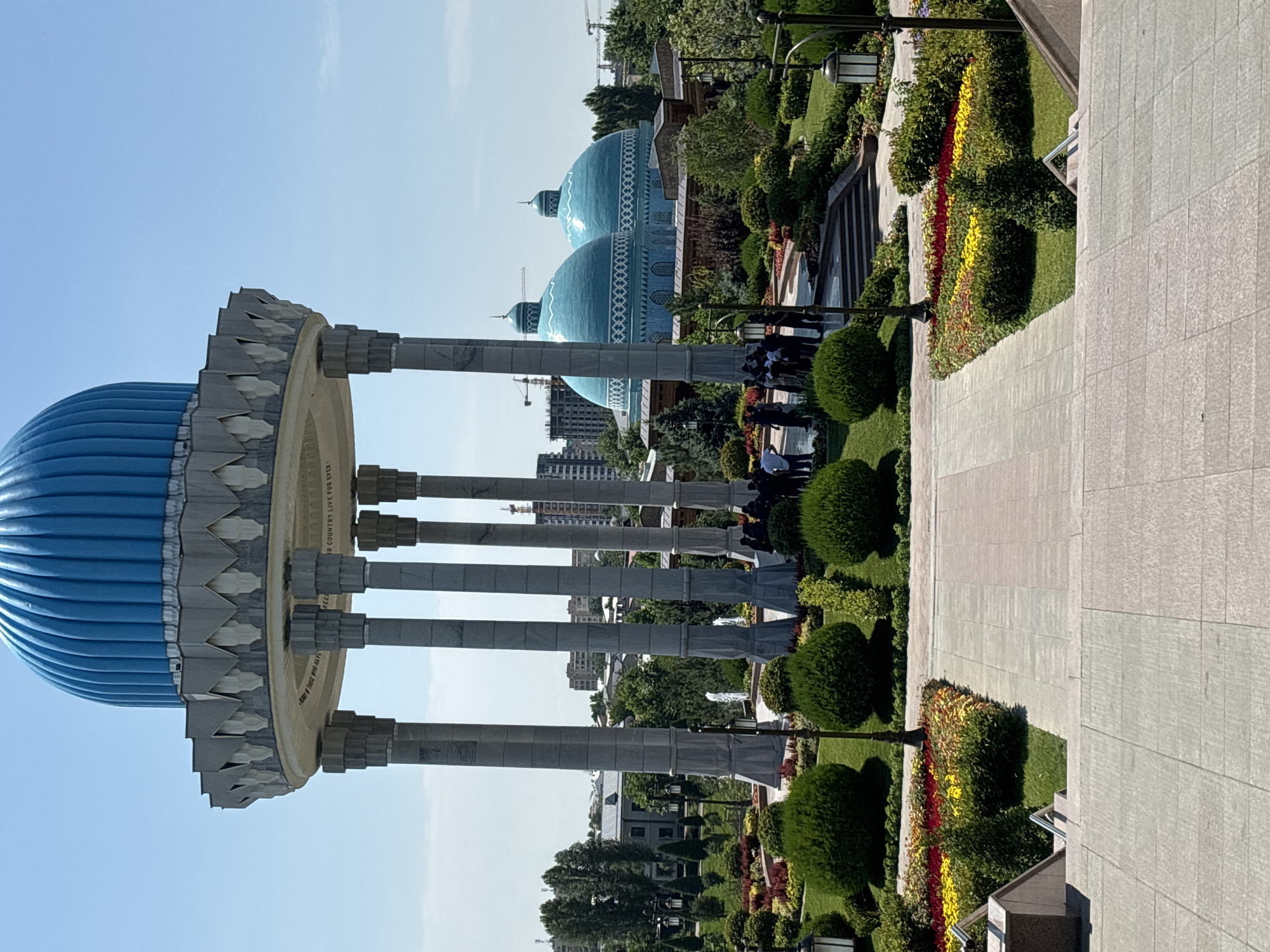 The image shows the Mausoleum of the First President of Uzbekistan, Islam Karimov, in the city of Tashkent, Uzbekistan. The mausoleum features a large blue dome and is surrounded by a manicured garden with colorful flower beds, trimmed bushes, and trees. In the background, two other blue domes are visible, along with modern buildings and construction cranes. A group of people are gathered in front of the mausoleum, seemingly observing the structure or posing for photos. The weather is clear and sunny with a blue sky. The foreground shows a wide stone staircase leading up to the garden area.