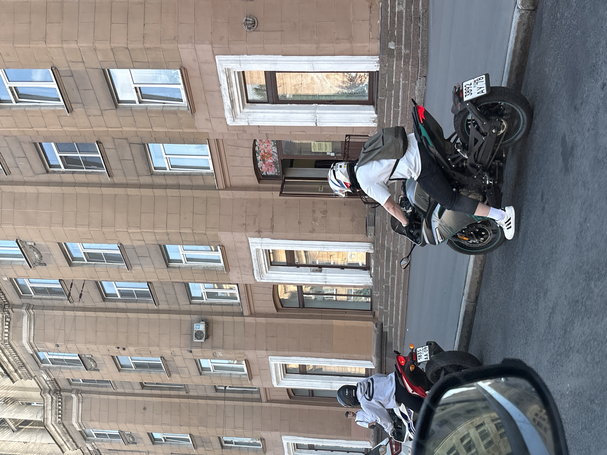A street in Saint Petersburg, Russia, features a person on a black and gray motorcycle bearing the license plate 3892 AY 78. The rider, wearing a white helmet, white shirt, black pants, white socks, and white Adidas sneakers, is positioned next to a tall, brown building with numerous windows. Another motorcycle, red and white, is visible in the background. The license plate of the second motorcycle is 1835 AA 09. A side mirror on the right provides a glimpse of the building reflected within.