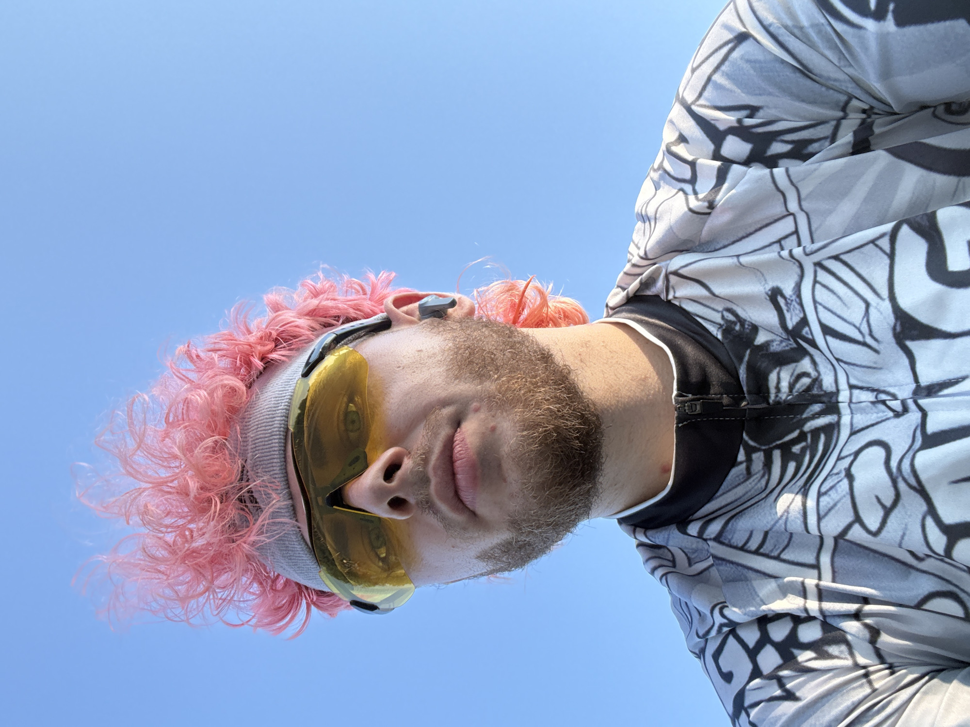 A person with pink hair and a beard is viewed from a low angle against a clear blue sky. They are wearing yellow-tinted goggles and a gray headband. The person is wearing a gray and white patterned athletic jacket with a black zipper. The location is not identifiable as Đà Nẵng, Vietnam.