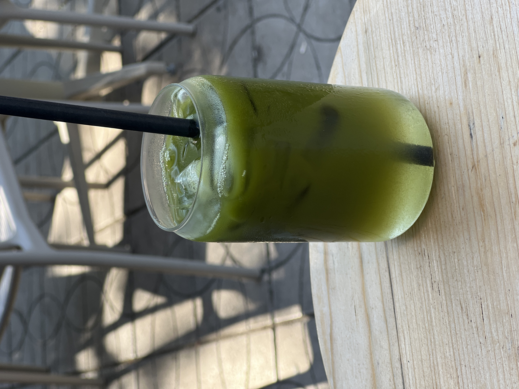 An iced matcha latte with a black straw sits in a clear glass on a light wooden table. The glass is covered in condensation, indicating it is a cold beverage. In the background, the blurred image of outdoor seating with white chairs and a patterned floor suggests a cafe or restaurant setting in the city of Bar, Montenegro. Sunlight casts shadows through the chairs and onto the floor.
