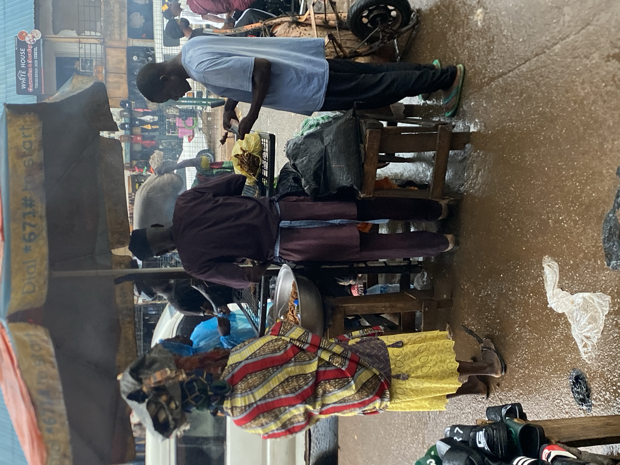In Bukuru, Nigeria, a street scene unfolds beneath an orange and yellow striped umbrella. Three individuals are the central focus. To the left, a person in a vibrant yellow and red striped outfit with a matching headscarf and a yellow skirt carries a large bundle on their head, partially obscured by a black and grey fabric covering. They stand near a metal basin filled with food. In the center, a person wearing a maroon tunic and trousers, cinched with a blue cloth belt, stands before a grill. To the right, a person in a light blue t-shirt and black trousers holds a piece of meat. The ground, a wet, brown surface, reflects the overcast sky and suggests recent rain. In the background, market stalls display various goods, including clothing. A white van is partially visible to the left, and a cart with a wheel is positioned behind the person in the blue shirt. The overall impression is of a bustling outdoor market, possibly after a rain shower, with vendors preparing and selling food.