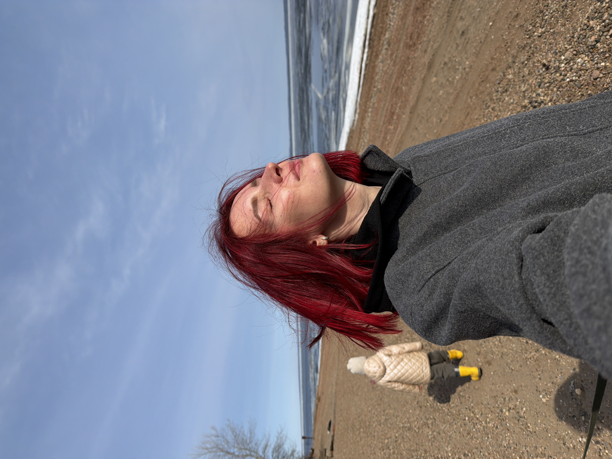 A person with red hair is seen looking upward against a blue sky. The individual is wearing a grey coat. Behind them, there is a body of water with ice on the edge, meeting a sandy beach. Another person, wearing a beige quilted jacket and yellow boots, is walking on the beach. A tree is visible in the lower left corner. The location is in the city of Chaikovsky, Russia.