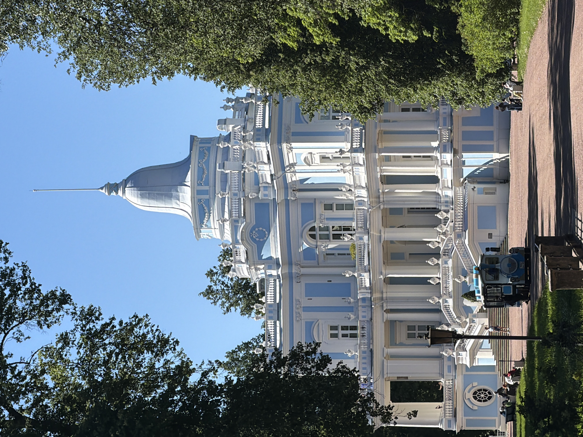 This is a vertical, eye-level shot of a grand, ornate building with blue and white accents, possibly a palace or historic structure, in a park-like setting. Lush green trees frame the top and sides of the image. The architecture features intricate white detailing, columns, balconies, and a prominent domed tower with a spire.
In the middle ground, a small blue train-like vehicle is parked on a pathway. Several people are visible, some sitting on benches, others walking along the path or near the building. The sky is a clear, bright blue, suggesting a sunny day. The shadows cast by the trees and the building indicate it is likely daytime, possibly late morning or afternoon. The overall atmosphere appears peaceful and leisurely, as people enjoy the outdoor environment and the impressive architecture. The scene evokes a sense of historical grandeur and natural beauty. No visible text is present in the image.