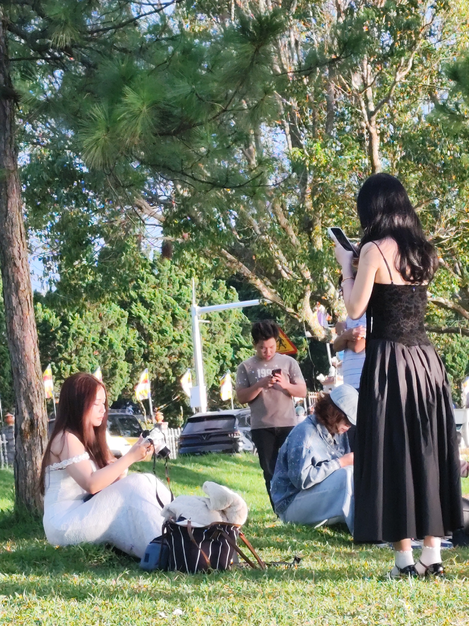 In Da Lat, Vietnam, a group of individuals are gathered outdoors on a grassy area, partially shaded by pine trees. In the foreground, a woman with long brown hair, wearing a white off-the-shoulder dress, sits leaning against a tree trunk. She is looking down at her phone. To her right, a black bag with white stripes and a blue pouch sits on the grass. Further back, a man with dark hair, wearing a grey t-shirt, stands and looks at his phone. To his right, a person wearing a denim jacket and light blue jeans squats, also looking at their phone. Behind them, a woman with long black hair, dressed in a black sleeveless dress with a patterned bodice and a full skirt, stands holding a phone. Her back is to the camera. In the background, there are vehicles and foliage, with flags visible on poles. The lighting suggests it is daytime.