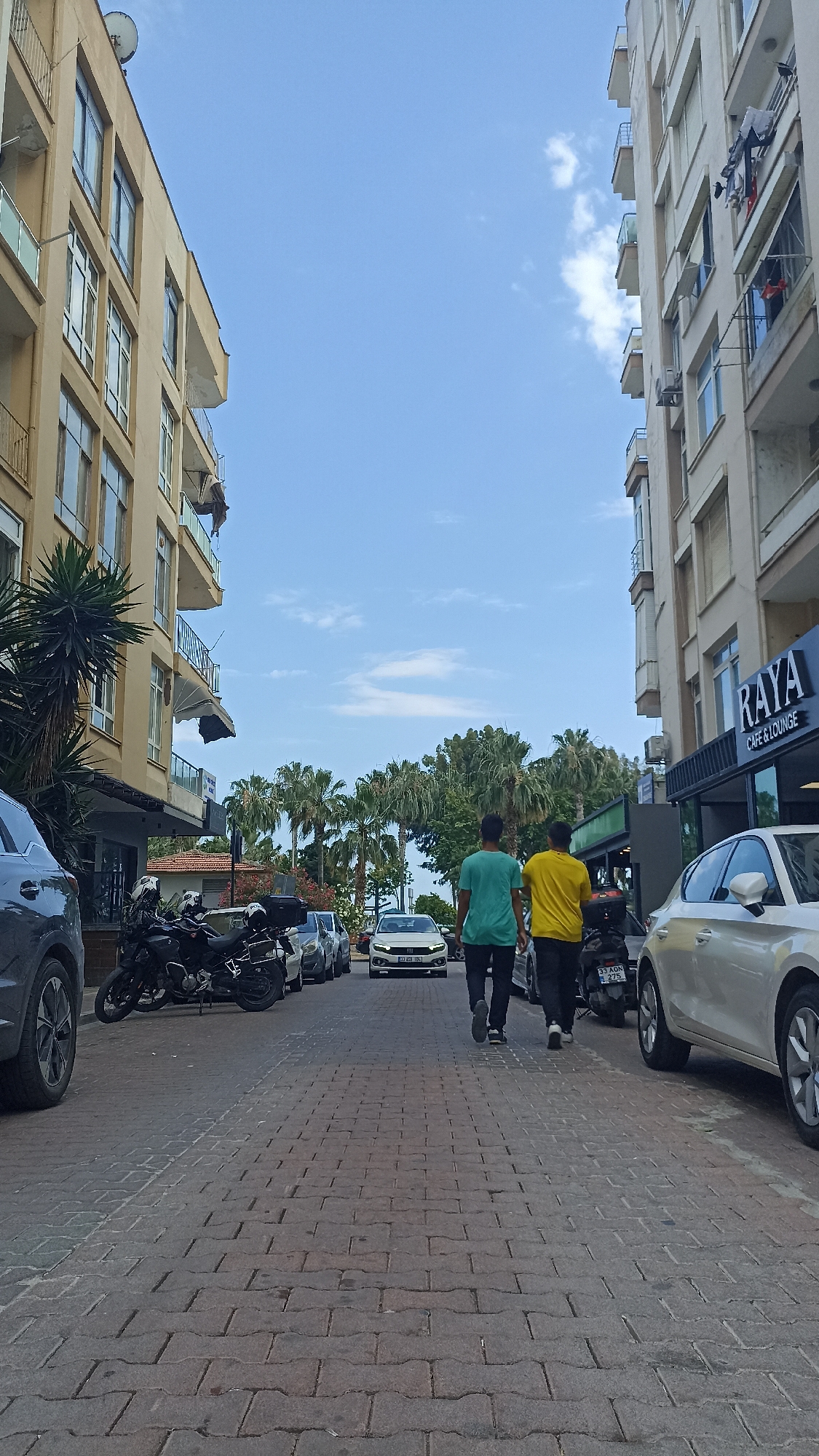 The image depicts a street scene in an urban environment. Two men, one in a turquoise t-shirt and the other in a yellow t-shirt, are walking away from the camera down a paved street. Several motorcycles are parked on the left side of the street. On the right side of the street, a building with a sign that reads "RAYA CAFE & LOUNGE" is visible, along with a parked silver car. Another building with balconies and windows is on the left side of the street, and a similar building is on the right. Palm trees and other greenery are present in the background. The sky is blue with scattered clouds. The overall scene suggests a street in a city, possibly in a warm climate.