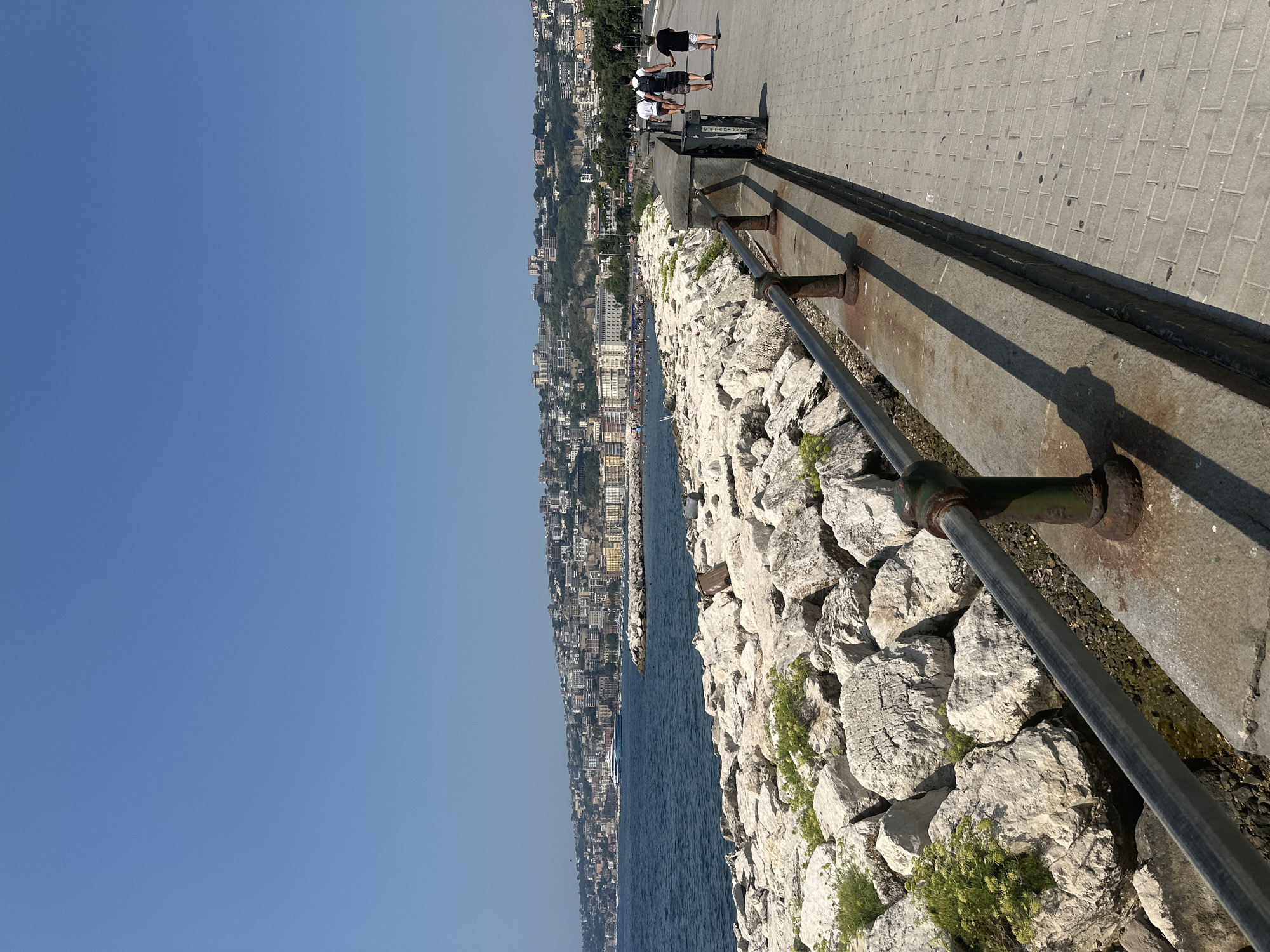 The image depicts a sunny day at a seaside promenade in Naples, Italy, likely along the Lungomare Caracciolo. The foreground features a walkway paved with light-colored rectangular stones, bordered by a low concrete wall and a metal railing with rusted supports. To the left of the walkway is a breakwater composed of large, irregular white rocks, with sparse green vegetation growing between them.
Beyond the breakwater, a deep blue sea stretches to the horizon, dotted with small boats and a prominent light blue yacht. The background showcases the cityscape of Naples, a dense collection of multi-story buildings in various shades of white, beige, and yellow, climbing up a hillside under a clear, bright blue sky.
Several people are visible walking along the promenade. In the mid-ground, on the right side of the walkway, a group of four individuals is walking away from the camera. The two adults in front are wearing white tops, and one is carrying a backpack. A child in a black shirt and shorts is walking behind them. Further in the distance, more people can be seen enjoying the seaside, some appearing to be on a beach area to the left. The overall atmosphere is one of a leisurely day by the sea.