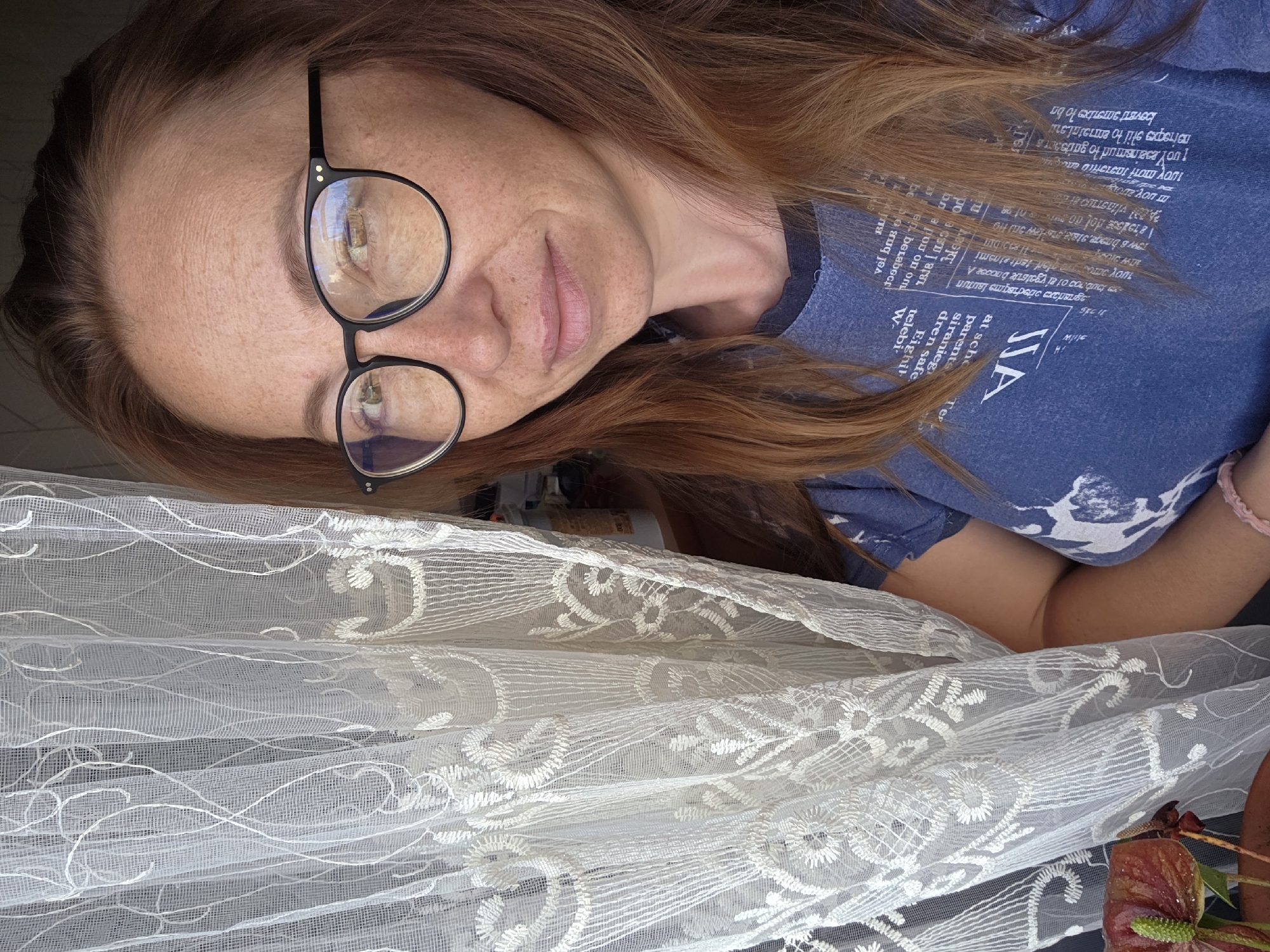 A medium shot shows a woman with shoulder-length auburn hair and fair skin with freckles, wearing black-rimmed glasses and a blue t-shirt with white text. She is standing near a white lace curtain with floral patterns. A red Anthurium flower is visible in the lower left corner of the frame. The background is blurred, with hints of shelves and items suggesting an indoor setting. The lighting appears to be natural, coming from the side. The woman's facial expression is neutral, and she is looking towards the camera. The scene appears to be in a residential or personal space. No specific location in Буренка, Russia, can be determined from the image.