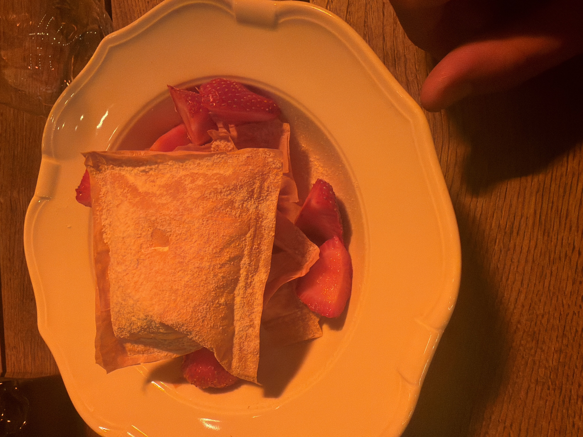 An overhead, close-up shot captures a dessert on a white, ornate plate, resting on a wooden surface. The scene is bathed in warm, dim, yellow-orange artificial light, suggesting an evening or indoor setting.
The dessert prominently features a light-colored pastry, likely a turnover or similar baked good, dusted generously with powdered sugar. Its surface appears flaky and slightly uneven. Arranged around and beneath the pastry are several bright red, sliced strawberries, glistening and appearing fresh.
The plate itself is white or off-white, with a gently scalloped or fluted rim, and has a slight sheen reflecting the warm light. To the top right of the plate, the tip of a human finger, possibly a thumb, is visible, partially obscured by shadow, suggesting interaction with the scene. A clear glass or drinking vessel, out of focus, is partially visible on the far left edge of the image. It has an etched or patterned surface, but no discernible text can be read. The background is a wooden table with a visible grain, providing a rustic contrast to the white plate. The image captures a moment before or during the enjoyment of the dessert.