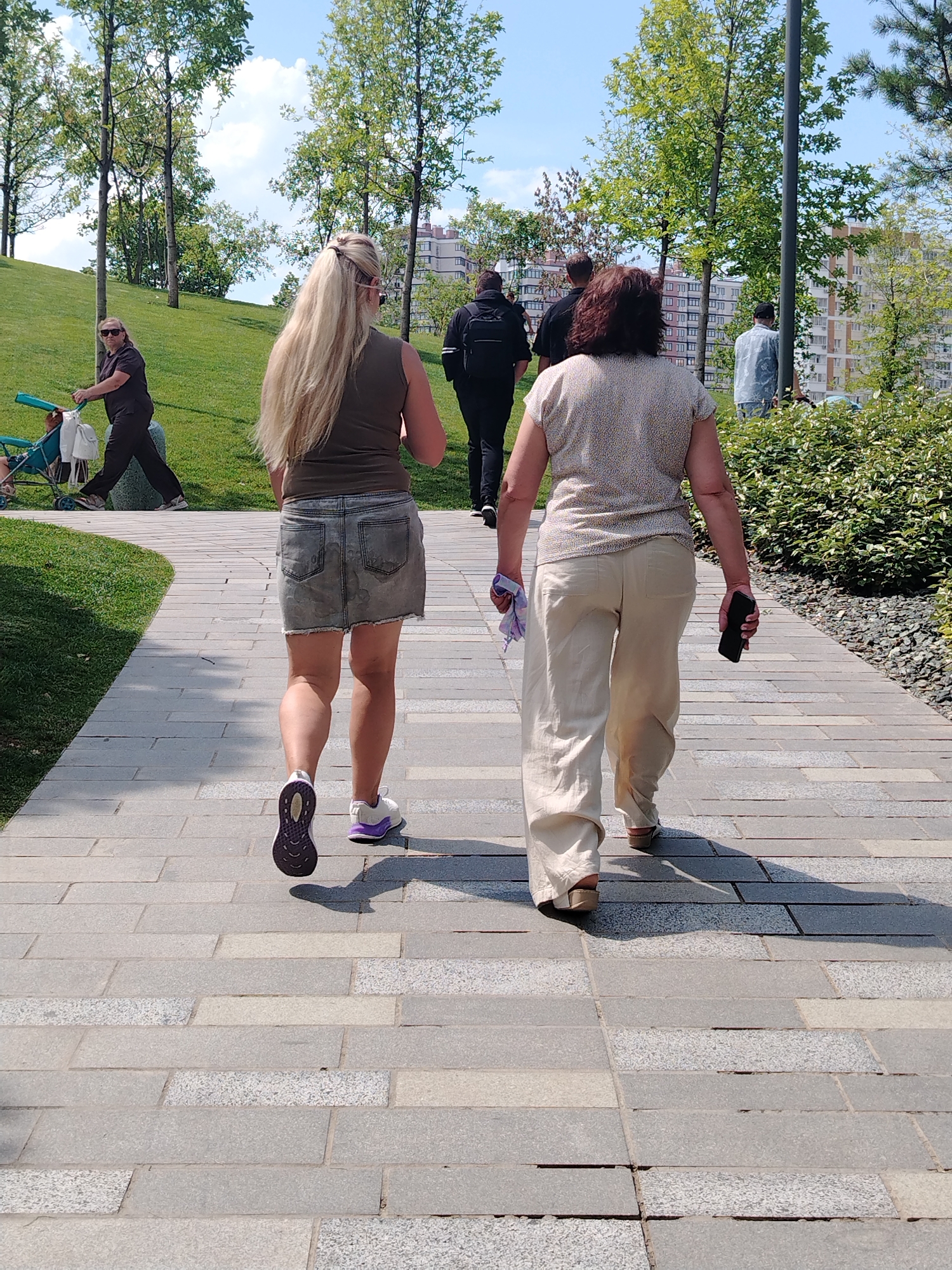 Two women are walking away from the camera on a paved pathway in a park. The woman on the left has long blonde hair and is wearing a dark tank top and a denim skirt. She has a purple and white sneaker on her left foot and a white sneaker with purple accents on her right foot. The woman on the right has dark hair and is wearing a patterned short-sleeved shirt and light-colored wide-leg pants. She is holding a black smartphone in her right hand and a purple and white cloth in her left hand.
In the background, there are trees, a grassy hill, and buildings. Other people are visible in the distance, including a woman pushing a stroller on the grassy area to the left. The scene appears to be in an urban park with well-maintained landscaping. The buildings in the background suggest a city environment, potentially in Krasnodar, Russia, given the context. The lighting indicates a sunny day.