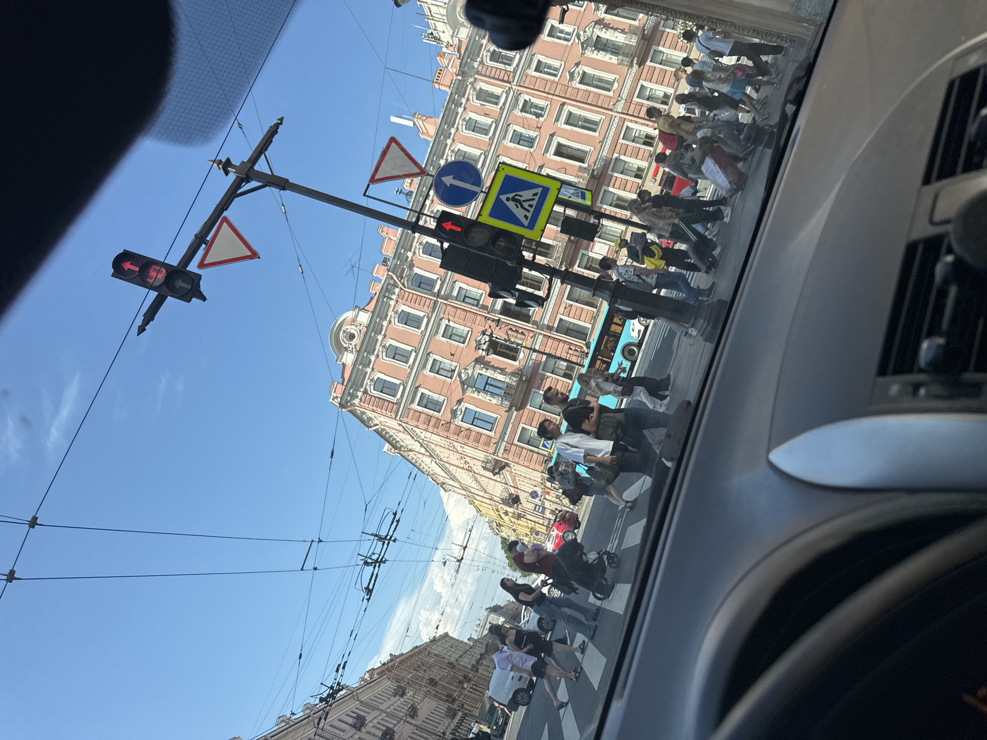 The image depicts a street scene in Saint Petersburg, Russia, likely taken from inside a vehicle. A group of pedestrians are crossing a street at a marked crosswalk. The traffic light shows a red signal for vehicular traffic and an upward arrow, indicating that straight-through traffic may proceed. The buildings on either side of the street are tall, with a traditional European architectural style, featuring ornate facades and multiple windows. Overhead, a complex network of tram or trolleybus wires is visible against a bright blue sky with some scattered clouds. The time of day appears to be daytime, with strong sunlight casting shadows. The foreground shows the interior of a car, including part of the dashboard and steering wheel, indicating the photographer's perspective.