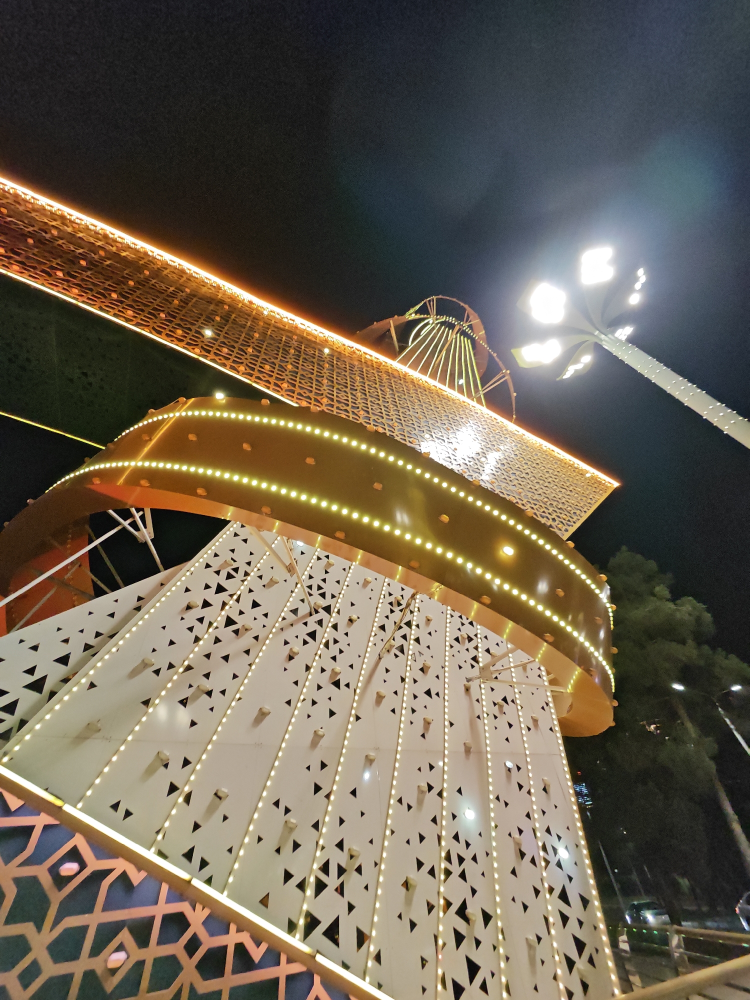 An extreme low-angle, wide shot captures a brightly illuminated modern architectural structure at night in Dushanbe, Tajikistan. The structure features white panels with triangular cutouts and is adorned with strings of small, warm-toned LED lights. A prominent golden, curved band wraps around the upper section of the structure, also outlined with LED lights. Above this, a latticed, golden-toned canopy with embedded lights extends towards the dark night sky. To the right, a tall, modern lamppost with multiple bright white lights illuminates the scene. In the background, silhouetted trees and distant lights suggest an urban environment. The overall impression is one of contemporary design and festive illumination, possibly part of a public space or an event.