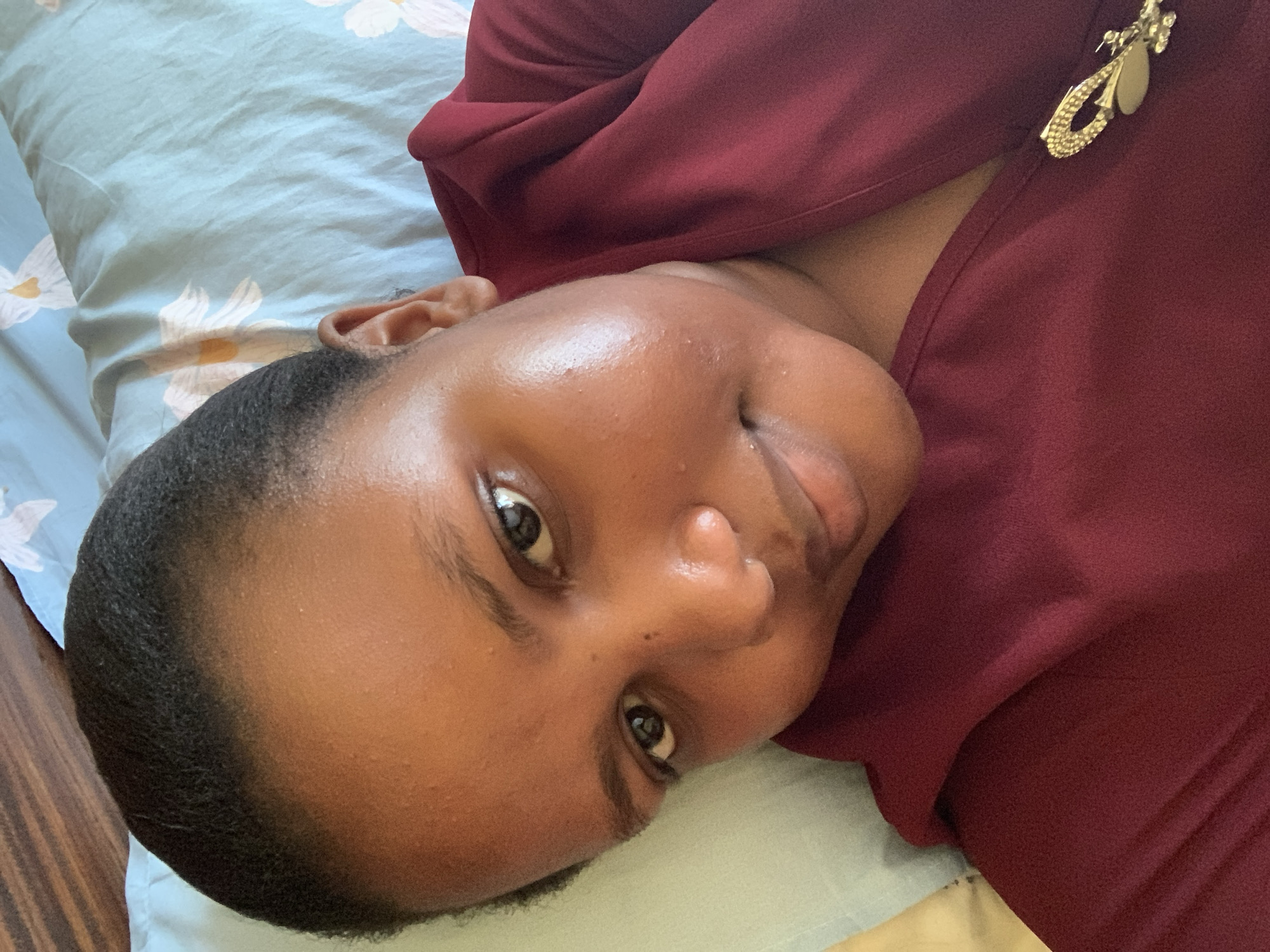 A close-up, tilted indoor shot features a woman with dark skin and short, dark hair, lying on her side. She is wearing a maroon-colored garment adorned with a golden decorative pin. Her eyes are open, and she is looking directly at the camera. The background consists of a light blue pillowcase with a floral pattern. The image does not provide enough information to definitively determine the location in Dar es-Salaam, Tanzania.
