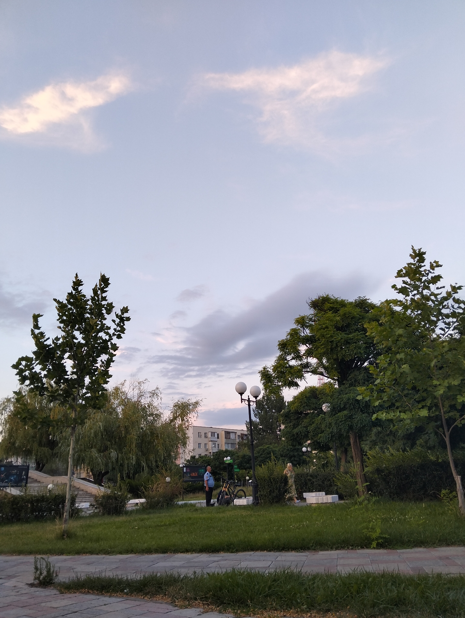 The image depicts a park scene in Astrakhan, Russia, during what appears to be the late afternoon or early evening. The sky is a pale blue with wispy, pale pink clouds, suggesting a sunset or sunrise. In the foreground, there is a paved walkway with visible grass growing between the pavers. Beyond the walkway, a well-maintained lawn extends into the park, dotted with various trees and shrubs.
To the left of the frame, there are stairs leading up to a level area with two large display boards, one showing images that appear to be space-related. A tall, thin tree with sparse, dark green foliage stands near these stairs. Further back, a weeping willow tree with cascading branches dominates the mid-ground.
In the center of the park, a man in a blue uniform shirt and dark pants stands facing left, with his hands clasped in front of him. Next to him, a bicycle is parked. Further to the right, another person, a woman in a light-colored patterned dress, is seen walking near a group of white, modular benches. A lamppost with two spherical lights is visible to the left of the man.
The background features a multi-story building, likely an apartment block, with visible windows and balconies. Dense green foliage from various trees and manicured bushes forms a backdrop for the park's activity. The overall impression is one of a public recreational space in an urban setting.