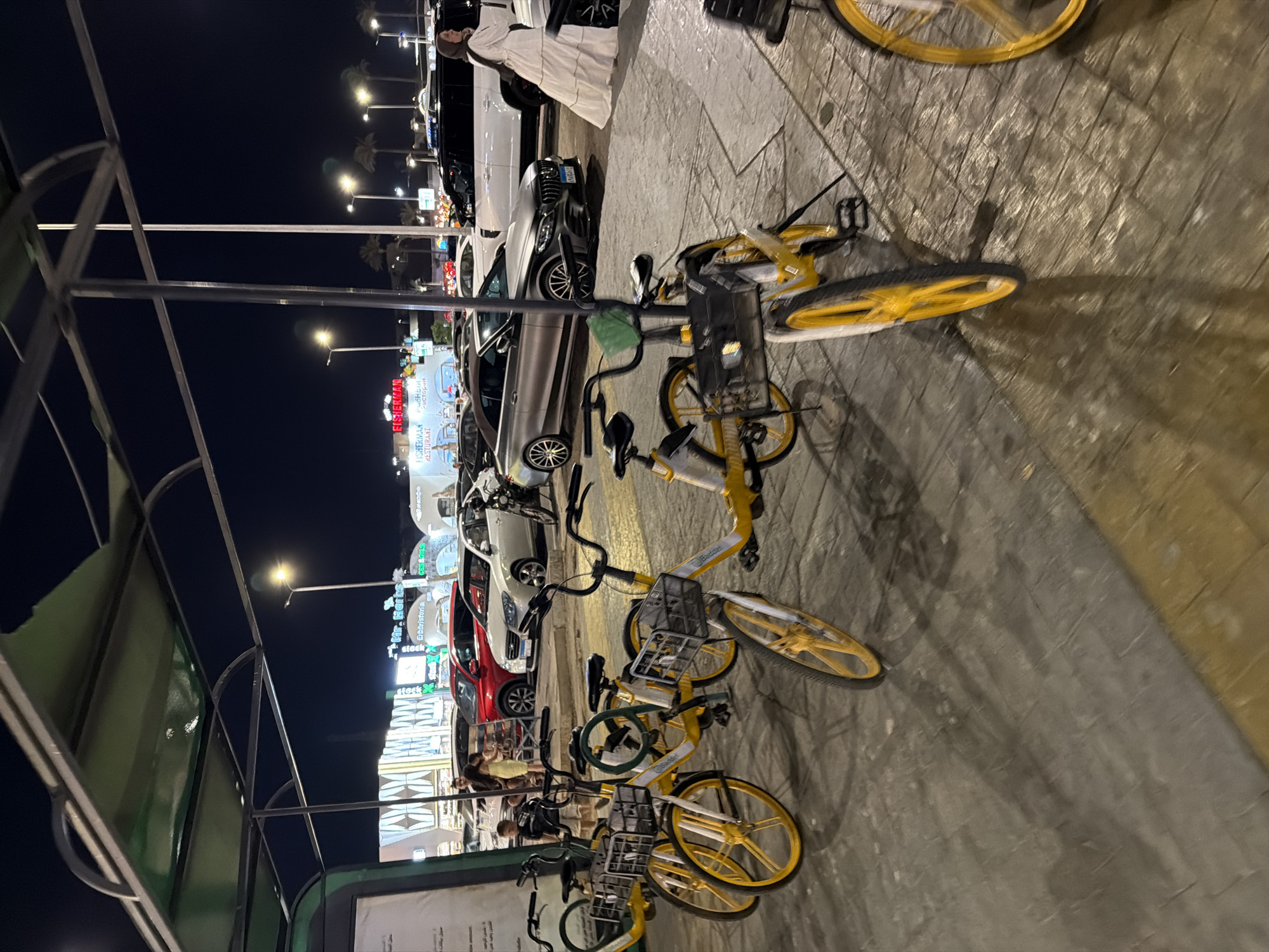 The image depicts a nighttime scene in Sharm El Sheikh, Egypt, likely at a promenade or entertainment district. In the foreground, several yellow rental bicycles are parked under a covered structure. These bicycles have baskets in front and are positioned on a paved surface. In the background, a variety of cars are parked, including a white SUV and a silver convertible. A woman in a long white dress is walking away from the camera on the right side of the image. Behind the parked cars, there are buildings with illuminated signs, one of which reads "FISHERMAN RESTURANT". Palm trees are visible in the distance, characteristic of the Egyptian landscape. The overall lighting suggests artificial illumination from streetlights and building lights, creating shadows on the ground. The scene appears to be a public area with a mix of recreational and commercial elements.