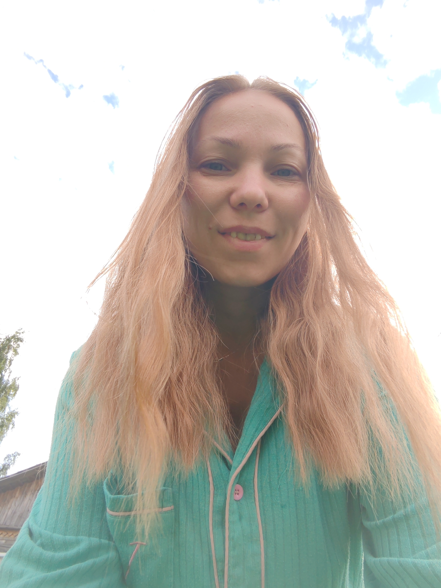The image shows a selfie of a woman with long, light blonde, wavy hair, taken outdoors with a bright, cloudy sky in the background. She is wearing a teal-colored pajama top with pink piping and a pink button. Her eyes are open and she is smiling faintly. To the left of the frame, a portion of a wooden building and some greenery are visible. The photograph appears to have been taken in a rural or suburban setting in the city of Chuchlem, Russia.