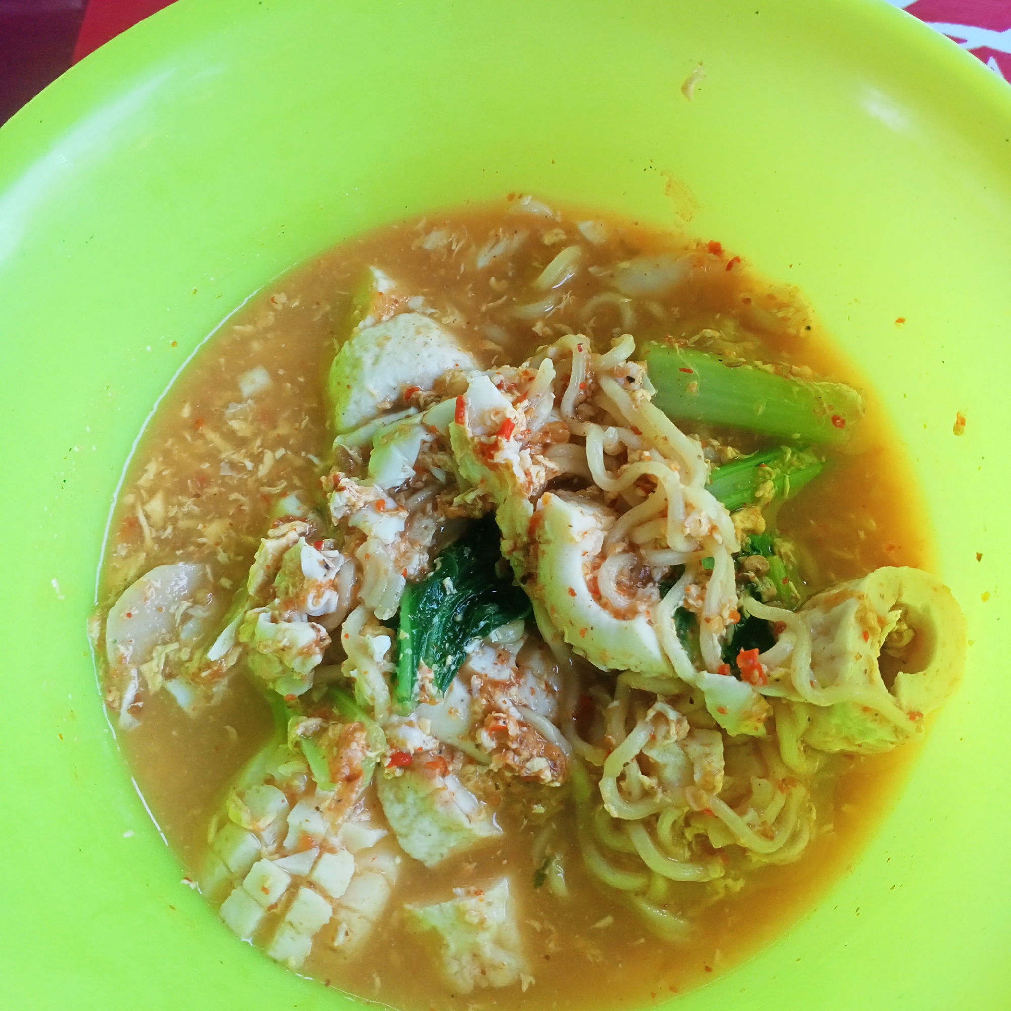 An overhead, close-up shot shows a bowl of food with a lime-green rim. The dish appears to be a noodle soup with various ingredients, including white noodles, chopped white and orange pieces, dark green leafy vegetables, and what looks like sliced white meat or seafood. The broth is a light orange-brown color and has a slightly frothy texture. Small red specks, likely chili flakes, are visible throughout the dish, suggesting it is spicy. The bowl is filled nearly to the brim with the food. The background is slightly blurred, but it appears to be a flat, red surface with a white and red striped pattern on the top right. There are no people visible in the image. The scene is a close-up of a food dish served in a bowl, likely in a restaurant or home setting in Kedung Dowo, Indonesia, given the context of the request.