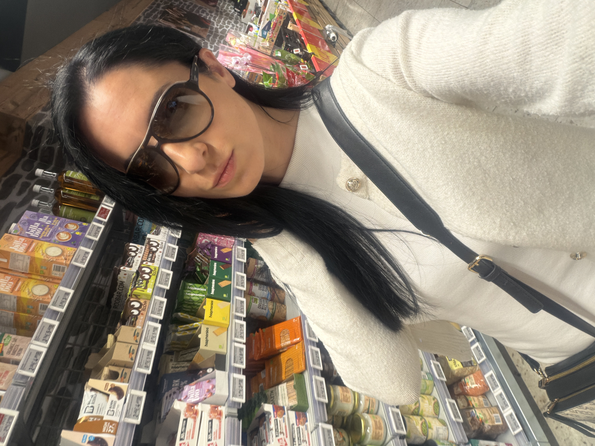 A woman is taking a selfie in a supermarket aisle. She is wearing large sunglasses and a white cardigan with a black shoulder bag. Shelves lined with packaged goods are visible behind her, including items labeled "billa bio" and "ROOBAR.COM," which suggests the location is a Billa supermarket. Some of the text on the labels are in a foreign language. The OCR data shows that the products are labelled "billa" and "ROOBAR" with the mention of "organic". Without further information, it is not possible to verify the exact location in Pleven, Bulgaria, only that the supermarket is a Billa branch.
