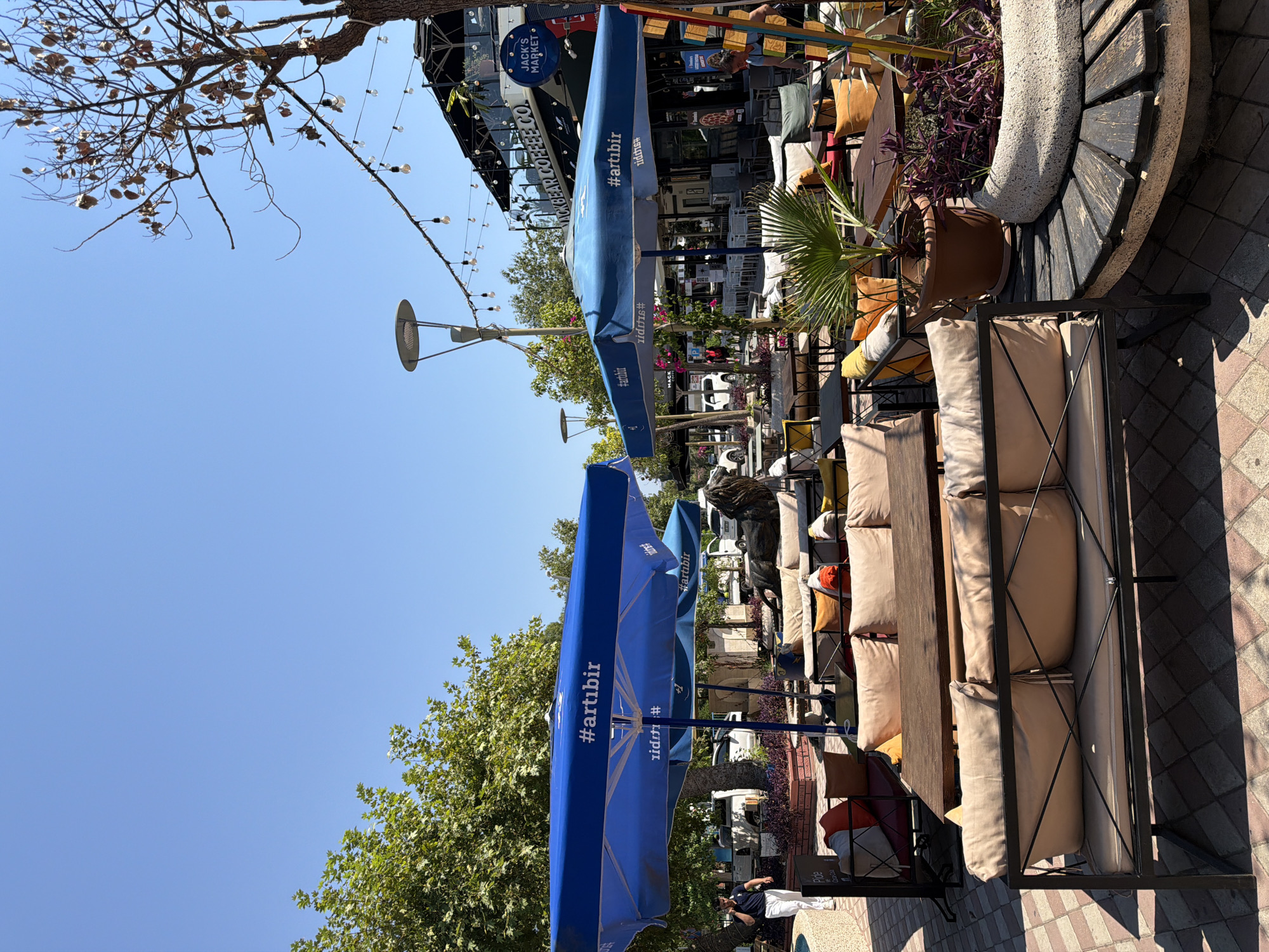 The image captures an outdoor seating area of a cafe in Kemer, Turkey. The scene is characterized by bright sunlight and a clear blue sky. In the foreground, a modern outdoor sofa with beige cushions is prominently featured, set against a patterned tiled ground. Several other tables and seating arrangements, including sofas and chairs with colorful cushions, are visible further into the scene, under blue umbrellas bearing the "#artibir" logo.
Behind the seating area, there is a building with signage for "JACK'S MARKET" and "BLACKBEAR COFFEE CO." String lights are suspended overhead, suggesting an evening ambiance despite the daytime lighting. Various trees and potted plants, including a palm tree, add greenery to the environment. In the background, a few people are present, including a man walking on the left and another person near the building on the right. Cars are parked along the street behind the cafe. A decorative lion statue is also visible. The overall impression is that of a bustling, well-maintained outdoor dining or relaxation space in a Turkish city.