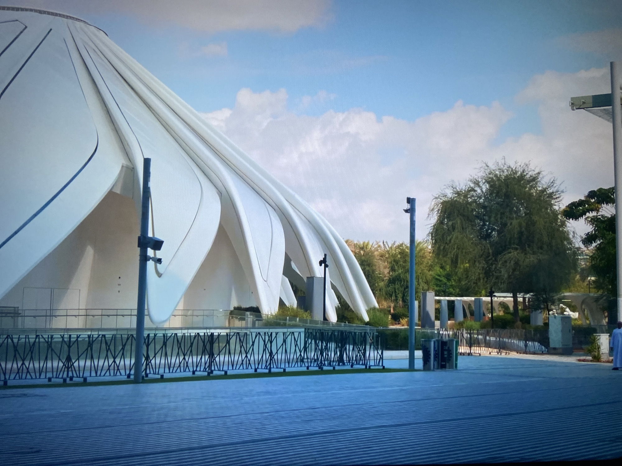 The image shows a modern architectural structure with a white, curved roof resembling petals, possibly part of a pavilion or exhibit. In the background, there are trees and park-like landscaping. A low, zigzagging fence runs along the front of the structure, with light poles interspersed. The foreground consists of a paved plaza with a linear pattern. A person in traditional white clothing stands to the right of the scene. The sky is partly cloudy. Location: Unknown, Unknown.