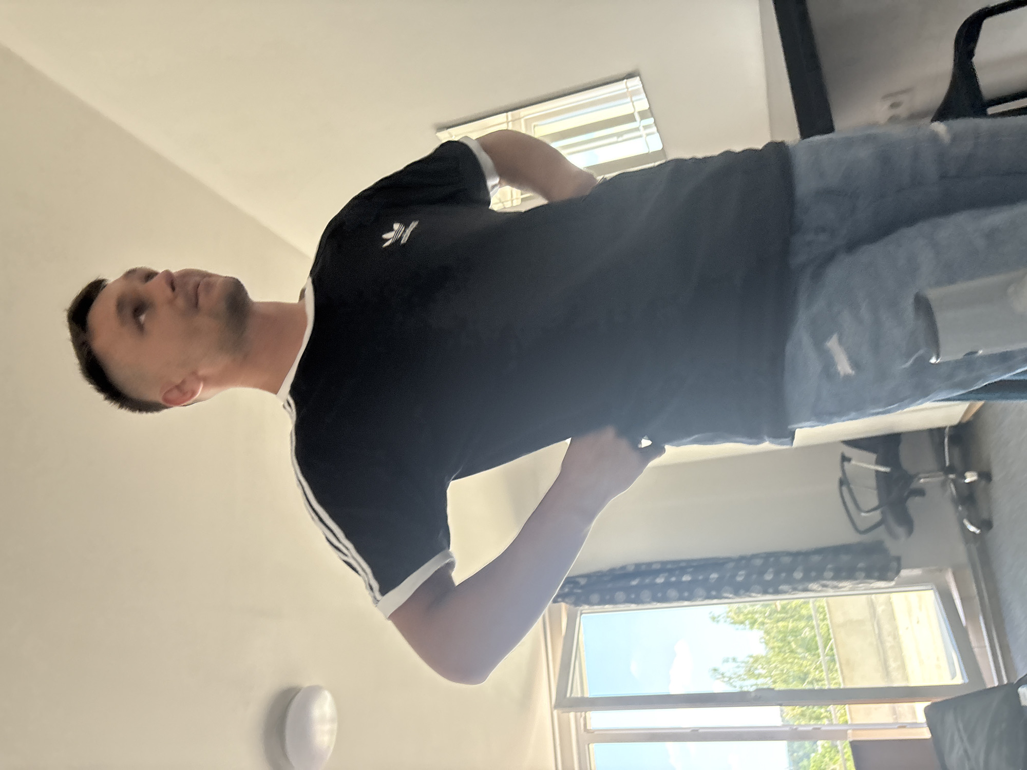 A young, fair-skinned man stands in what appears to be a bedroom, looking to his right. He is wearing a black Adidas t-shirt with white stripes on the sleeves and light blue ripped jeans. His hands are on his hips. Behind him, to the left, is a large window that opens onto a balcony with greenery visible outside. The room has white walls and a ceiling light. To the right, a black desk and a chair are partially visible. The lighting suggests daytime.