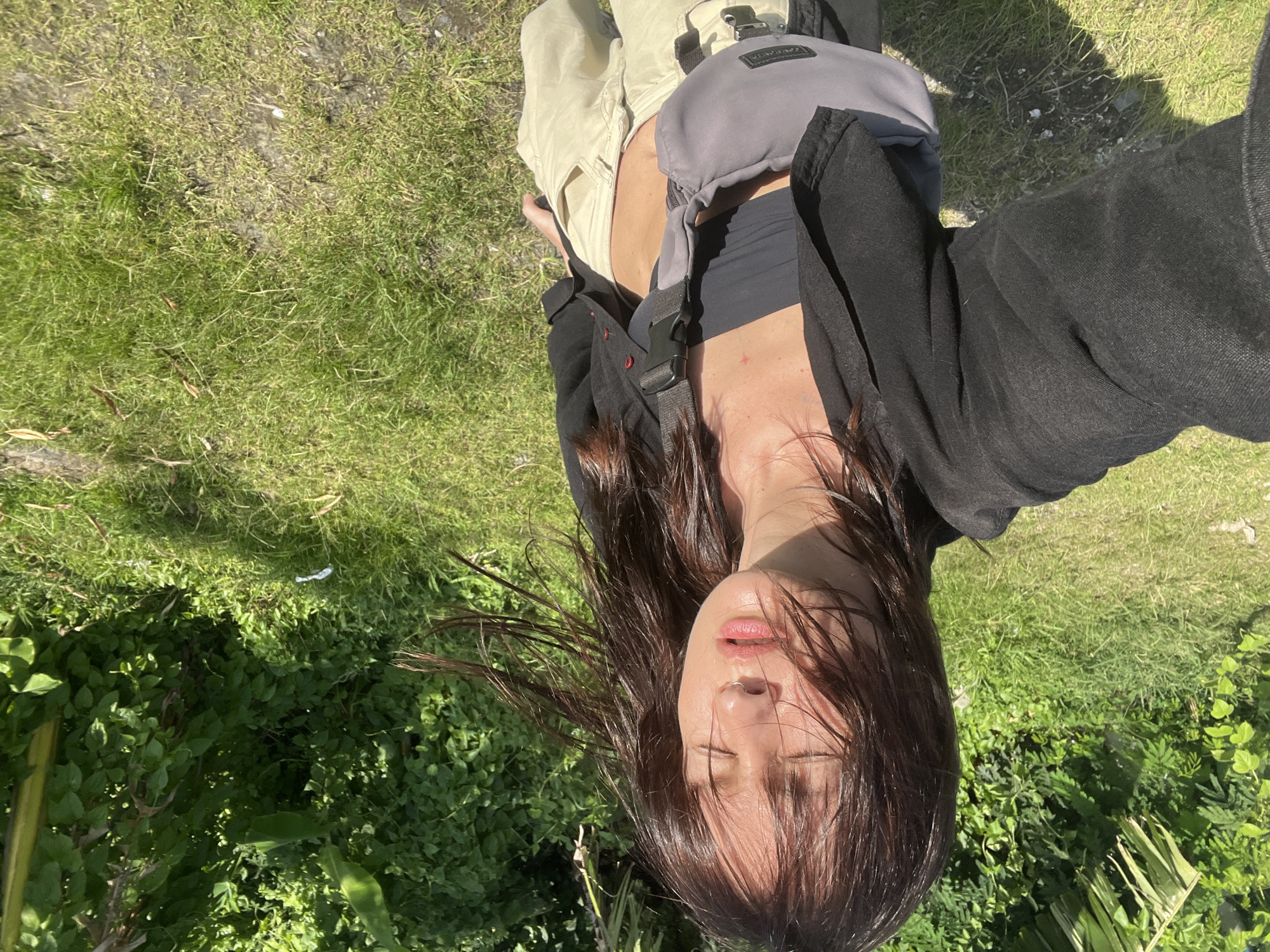 A selfie photograph features a woman with long, dark hair, closed eyes, and a nose ring. She is wearing a black top and jacket, with a grey sling bag across her chest. She appears to be outdoors, lying on a combination of grass and leafy vegetation. Part of her khaki pants are visible on the left side of the image. The background consists of greenery, with some patches of grass. The photograph was likely taken in North Kuta, Indonesia.