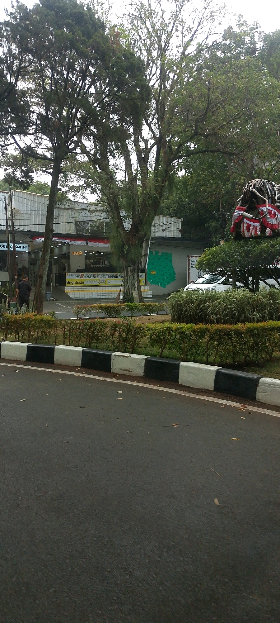 This is a medium-distance, eye-level shot of a street scene in Bandung, Indonesia, featuring a building with storefronts, trees, and a statue. The building, likely a commercial establishment, has a white facade with large windows and signage that includes "HOUS smith" and "RUSS". A yellow and white tiled facade runs along the lower part of the building, with a banner that reads "Brightside" and displays operating hours from 19:00-21:00. To the left of the building, a person in dark clothing is walking away from the camera. Several trees are prominent, with one large tree with textured bark directly in the center of the frame. To the right, a unique statue resembling a head with dreadlocks, adorned with red and white fabric, is visible atop a green bush. The foreground shows a paved road with a curb painted with alternating black and white blocks. A low hedge runs along the edge of the road. The sky is overcast, suggesting a cloudy day.
