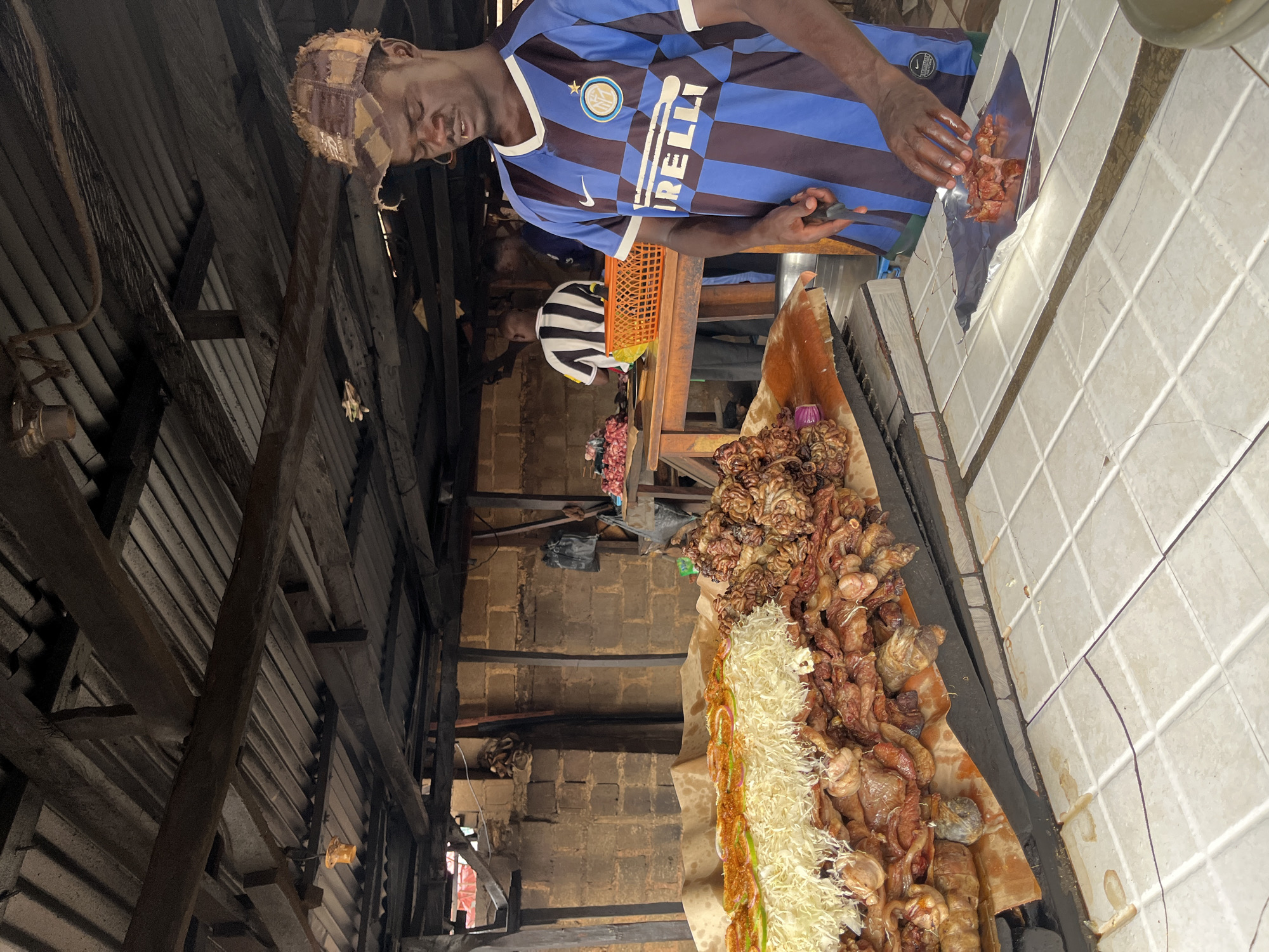 A man in a blue and black Inter Milan jersey stands behind a table laden with cooked meats and shredded cabbage, possibly in Bar Jirgi Summa, Nigeria. He is using a knife to portion meat onto foil. Other people stand behind him. The structure around him is made of brick and wood.
