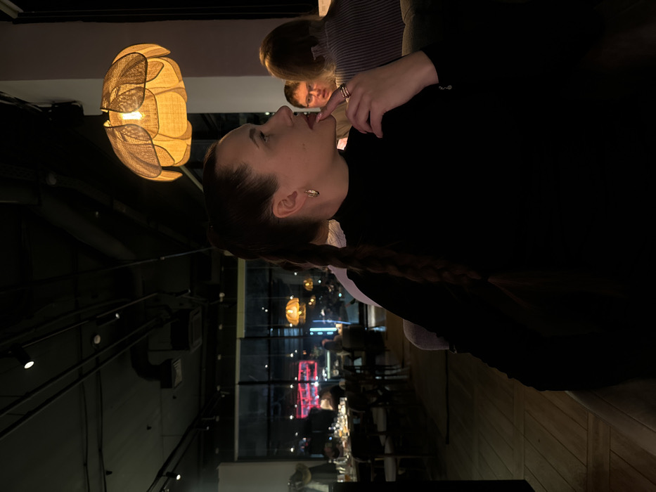 A woman with long, braided hair sits at a restaurant. She has her finger to her lips, lost in thought. A blurred figure sits to her right, and lights shine in the background. A lamp hangs in the foreground. The interior appears modern with dark walls and visible fixtures. The overall ambiance is dark and intimate.