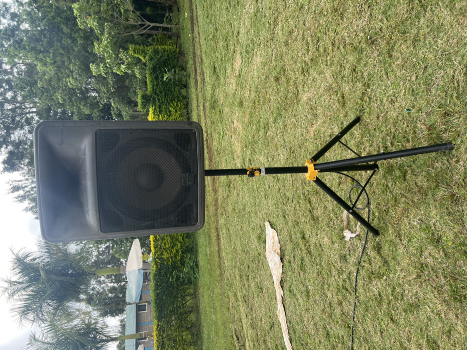 An outdoor scene in Makuyu, Kenya, features a black PA speaker mounted on a tripod stand, positioned on a vibrant green grass lawn. The speaker, facing left in the image, has a protective metal grille covering its cone and horn, with a faint logo visible on the grille. The black tripod speaker stand has bright yellow accents at its adjustable joint. A black power or audio cable snakes across the grass from the base of the stand.
In the mid-ground and background, dense green foliage and tall trees create a natural setting. Among the trees, a few palm trees are distinctly visible, and a light-colored building with a roof is partially seen behind some hedges. A long, light-colored piece of fabric or paper is lying crumpled on the grass near the speaker stand.
The lighting suggests it is daytime, possibly with an overcast sky, as there are no strong shadows. The overall impression is that an audio system is being set up for an outdoor event or gathering in a garden or park-like environment.