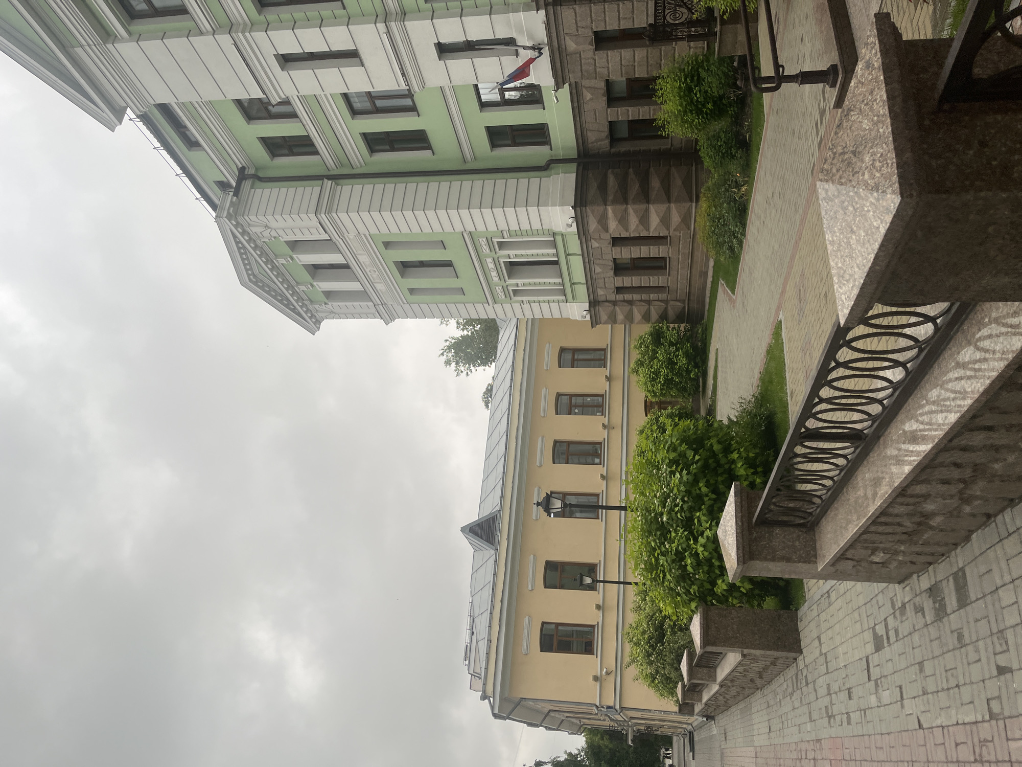 The image shows a view of two buildings on a cloudy day. The building on the right is tall and light green with white trim and a stone base. The building on the left is shorter, yellow, and appears to be older. Both buildings have multiple windows. In the foreground, there is a paved walkway with a stone and metal fence. Lush green bushes line the walkway and the buildings. A flag is visible on the right building. There are no people in the image.