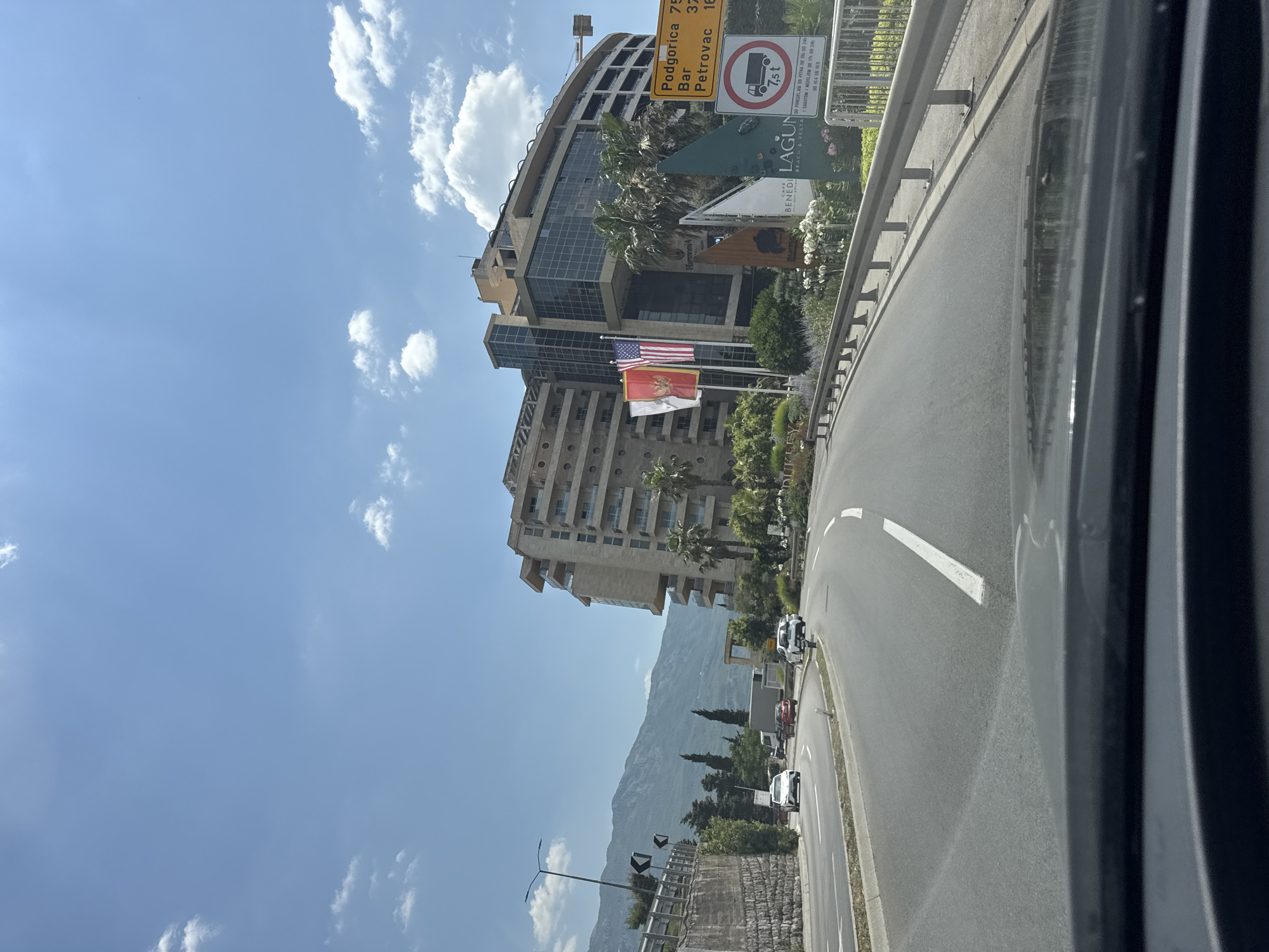 The image shows a view from a vehicle driving on a road in Budva, Montenegro, with a large, modern building on the right side of the road. The building appears to be a hotel or resort, with multiple balconies and a curved facade. Several flags are displayed in front of the building, including the flag of the United States, the flag of Montenegro, and another unidentifiable flag. Palm trees and lush vegetation surround the building. A road sign on the right indicates destinations such as Podgorica, Bar, and Petrovac, and also displays a truck weight limit of 7.5 tons. Several cars are visible on the road, and mountains form the backdrop to the scene. The sky is blue with scattered white clouds.