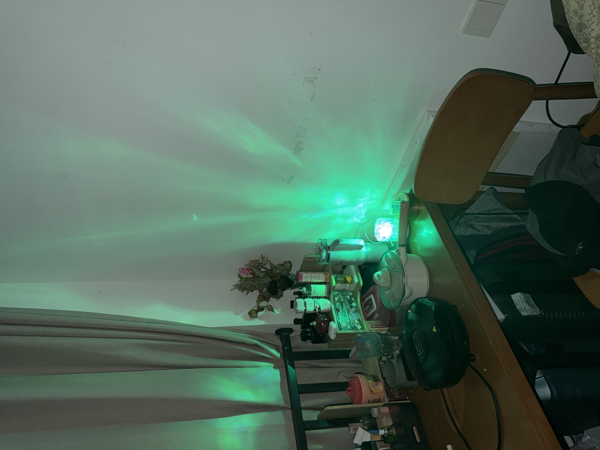 A low-angle, indoor shot of a cluttered shelf unit, illuminated with green light, positioned against a pale wall. The shelf is filled with an assortment of items including bottles, containers, a small bouquet of dried flowers, cookware, and various personal care products. A dark-colored curtain is visible to the left of the shelf. To the right, a wooden chair is positioned next to the shelf, partially obscured by bags and other items. The green light emanates from a source atop the shelf, casting patterns on the wall and curtain. The image is taken in Jiangning District, China.