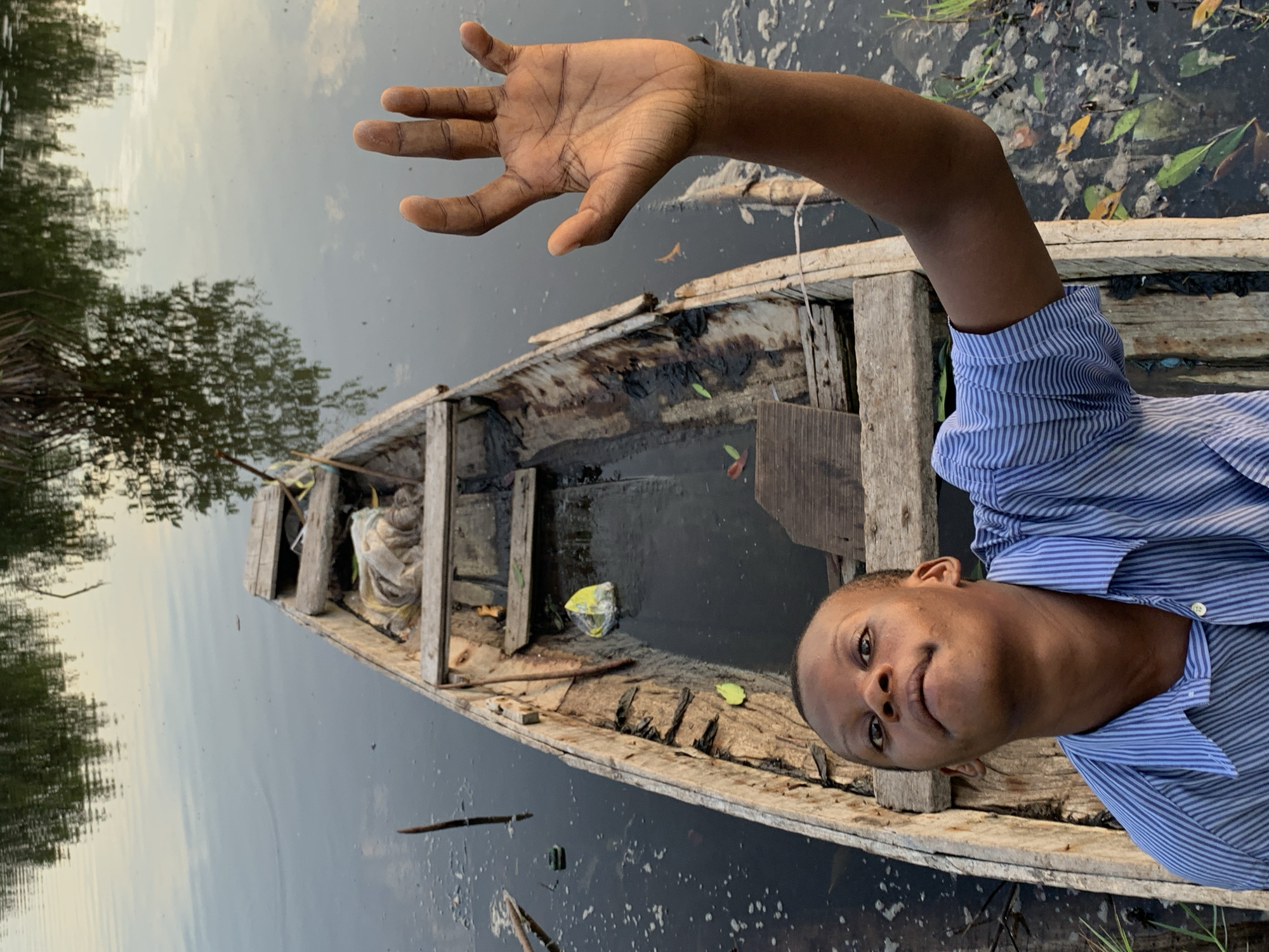 A close-up, portrait-oriented shot features a young person in a rustic wooden boat on dark, still water. The individual, appearing male with dark skin and a short buzz cut, occupies the right side of the frame, looking directly at the camera with a neutral to slightly curious expression. He wears a blue and white striped long-sleeved shirt. His right arm is extended upwards and to the left, with an open hand and spread fingers, possibly in a gesture or greeting.
The boat is a small, weathered wooden canoe or dugout, appearing aged with visible timber and some interior debris, including leaves and a yellow plastic bag, partially submerged in murky water collected within. The dark water fills the left and bottom sections of the frame, reflecting a light, overcast sky and the silhouettes of distant trees, indicating a natural, outdoor setting. Some small floating debris and scattered leaves are visible on the water's surface.
The soft, diffused lighting suggests an overcast day, early morning, or late afternoon. The scene evokes a tranquil moment in a riverine or delta environment, consistent with the location context of Ibasa, Nigeria. There is no visible text in the image.