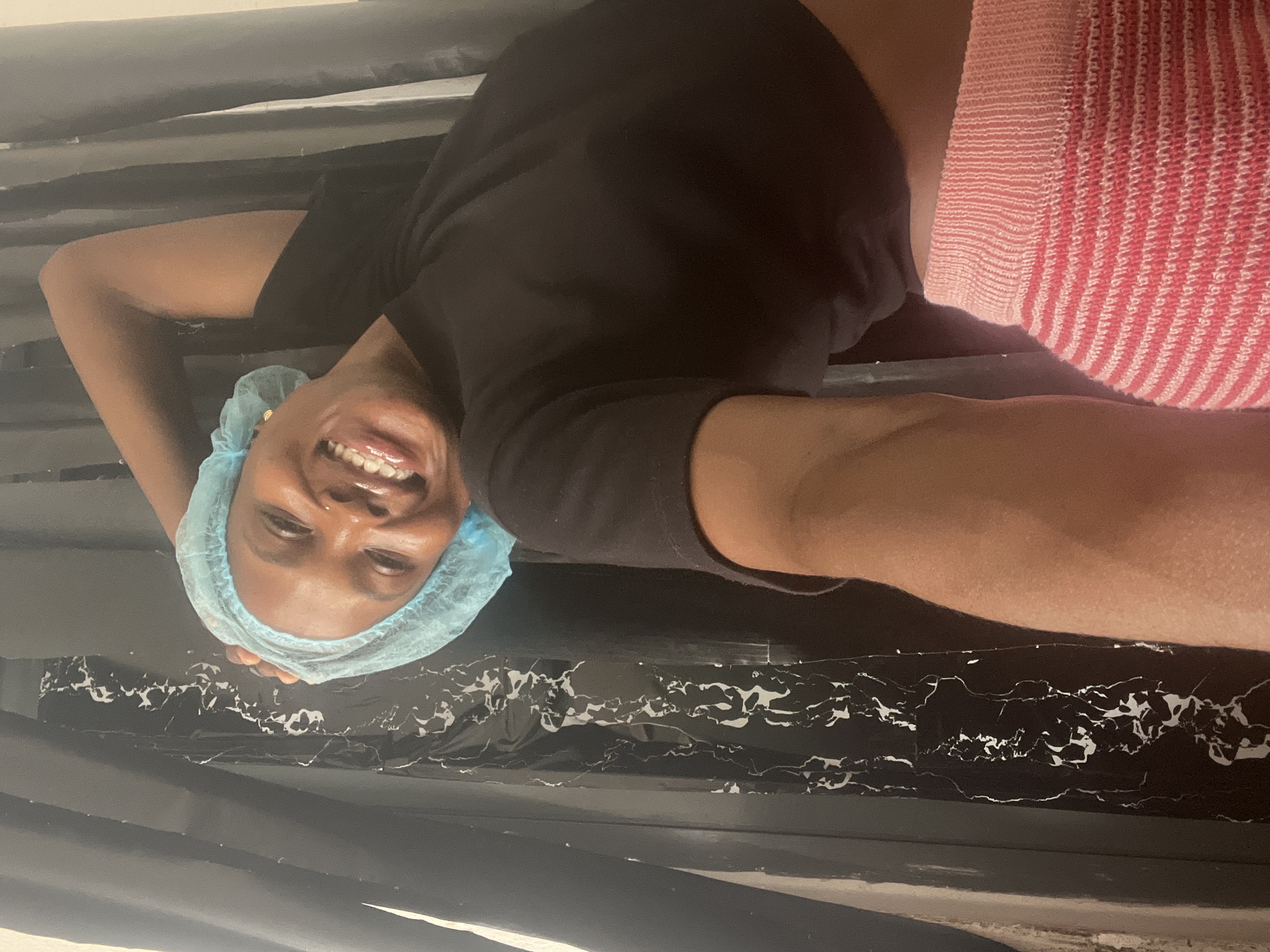 A smiling woman wearing a blue surgical cap and a black top is pictured in what appears to be an indoor setting. The background features black and white marble-like patterned surfaces, suggesting possible modern interior design. The location is in the city of Lekki, Nigeria. The woman's left arm is visible and extending forward towards the camera. There is also a partially visible striped object, possibly clothing or a towel, in shades of pink and white.