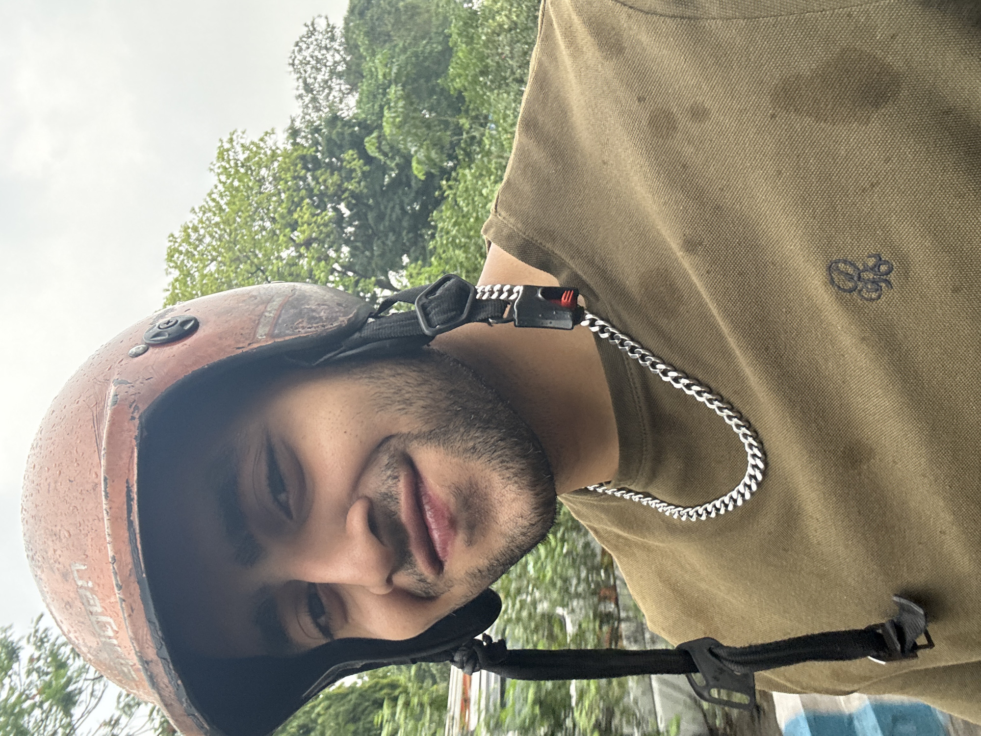 A medium, eye-level shot depicts a person with medium skin tone, dark hair, and a trimmed beard wearing a brown helmet, a silver chain necklace, and a tan, short-sleeved shirt with a small embroidered design on the upper right chest. The helmet strap is visible and secured. The person is looking at the camera with a slight smile.
The background consists of green trees. It is daytime, and the lighting suggests a possibly overcast day. Given the visual cues, including weather and attire, it could be assumed that the location is Kolkata, India.
