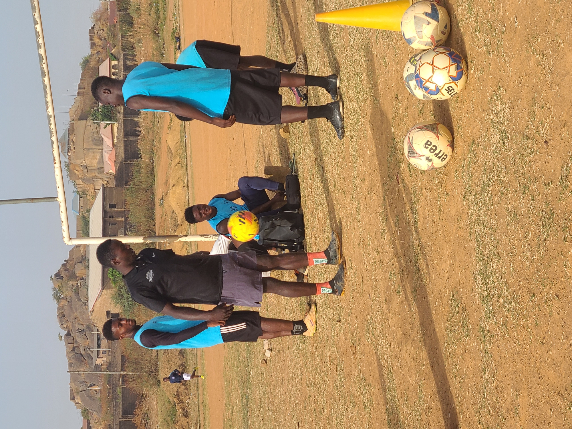 The image depicts a soccer field in Barikin Jirga Summa, Nigeria, with a group of people engaged in what appears to be a training session. Three soccer balls and a yellow cone are grouped together on the grass. Four individuals are standing, wearing blue and black soccer jerseys and shorts, with soccer cleats on their feet. A fifth person is seated, holding a soccer ball. In the background are buildings and rocky formations, typical of the area. A soccer goalpost is visible on the left edge of the frame. Another figure is walking on the field in the distance. The ground appears to be a mix of dirt and sparse grass.