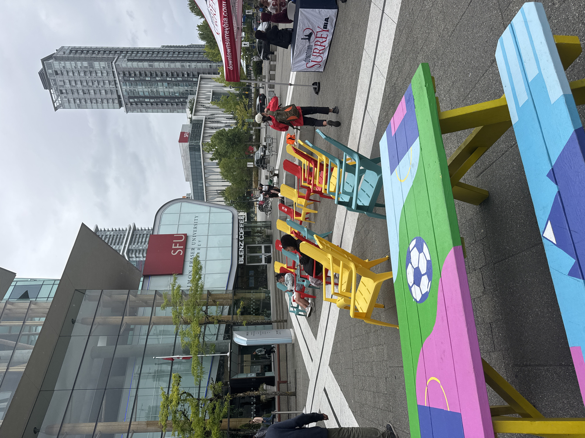 The image captures an outdoor public space in Surrey, Canada, likely near Simon Fraser University (SFU) as indicated by signage. The scene is set on a cloudy day, with a modern architectural backdrop featuring a glass-faced building with the SFU logo and "Simon Fraser University" visible. In the foreground, a vibrantly painted picnic table, adorned with a soccer ball motif and geometric shapes in green, pink, blue, and yellow, is prominent. Surrounding the table are several colorful Adirondack chairs in red, yellow, and teal, some of which are occupied. Two individuals are seated in red chairs, one looking at a phone, while another person in a red jacket and backpack walks away from the camera. In the background, a tall residential skyscraper stands against the overcast sky, and a red and white tent with "downtownsurreybia.com" and a "Downtown Surrey BIA" logo suggests a local business improvement association event or presence. Other individuals are seen in the distance, and some bicycles are parked nearby. The ground is paved with interlocking stones and marked with white lines, creating a geometric pattern.