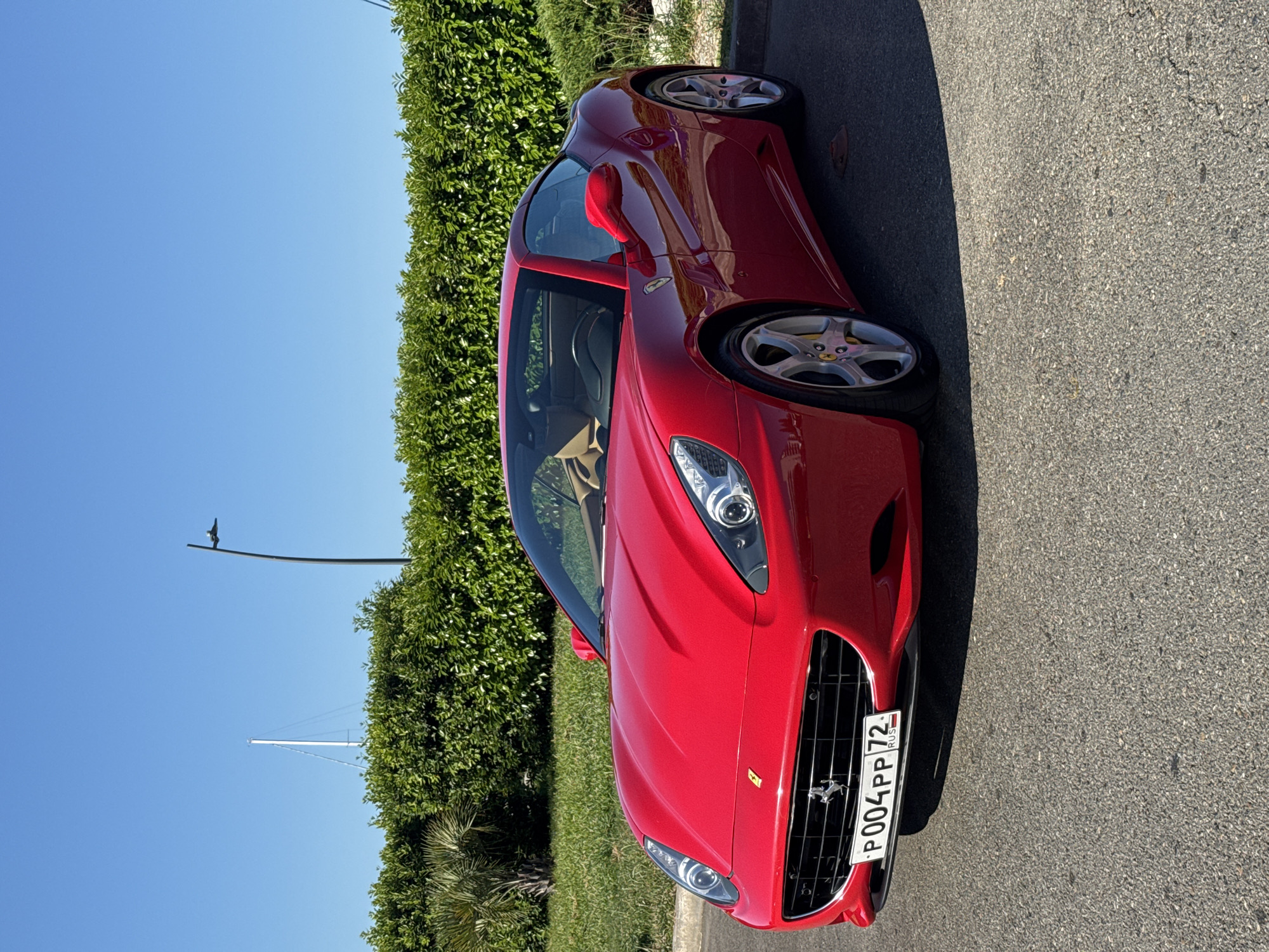 A red Ferrari California is parked on a paved surface in front of a manicured green hedge under a clear blue sky. The car is angled slightly towards the camera, showcasing its front and right side. A license plate with the inscription "P004PP 72 RUS" is visible on the front of the vehicle. In the background, a tall white mast, possibly from a sailboat, is visible above the hedge, along with a modern street lamp. The environment suggests a sunny day in a well-maintained outdoor area, potentially a promenade or a private driveway in Sirius, Russia. There are no people or animals present in the image.
