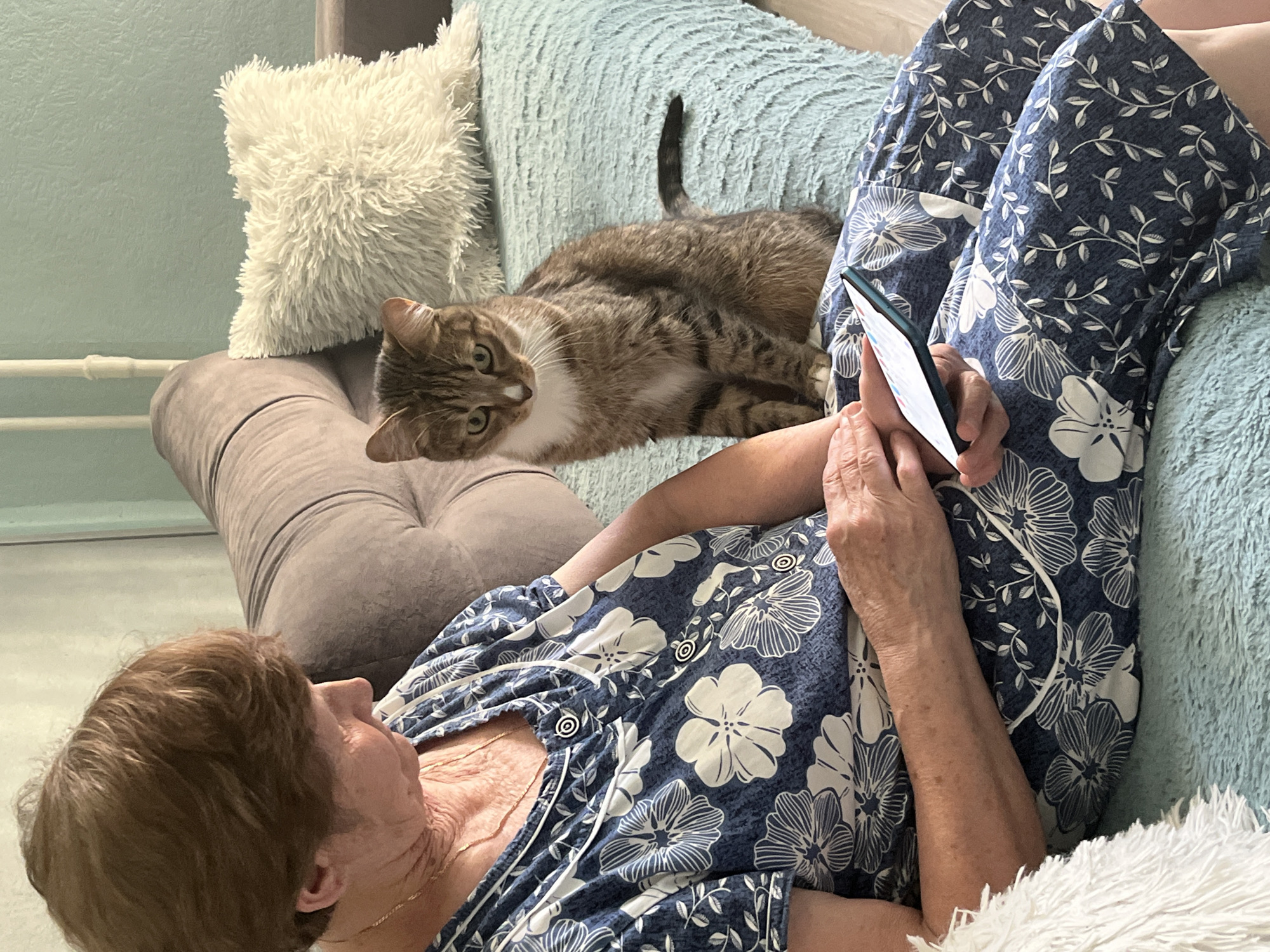 A woman is lying on a couch, looking at a smartphone. She is wearing a blue and white floral pattern shirt and pajama pants. A cat is lying on the couch beside her, looking at the camera. The couch is adorned with several pillows, including one that is fuzzy and white, and another that is blue and textured. The background is a light green wall. The location is unknown and there is no mention of Izhevsk, Russia.