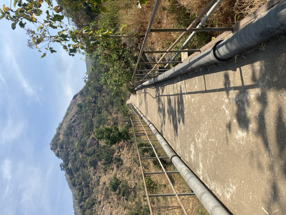 A concrete elevated pathway, approximately 3 meters wide, extends curvilinearly through a mountainous, vegetated landscape in Kpak, Nigeria, under a clear blue sky. The path is bordered on both sides by metal railings, each comprising two horizontal bars supported by vertical posts. Immediately adjacent to the outer base of these railings, thick, dark-grey utility pipes run parallel to the path. Sunlight casts distinct shadows of the railings and overhead foliage onto the concrete surface. The terrain flanking the pathway features dry, light-brown grasses and shrubs interspersed with green trees and visible rock formations. In the background, a large, rocky mountain adorned with patches of green vegetation rises prominently. No human presence or activity is observed.