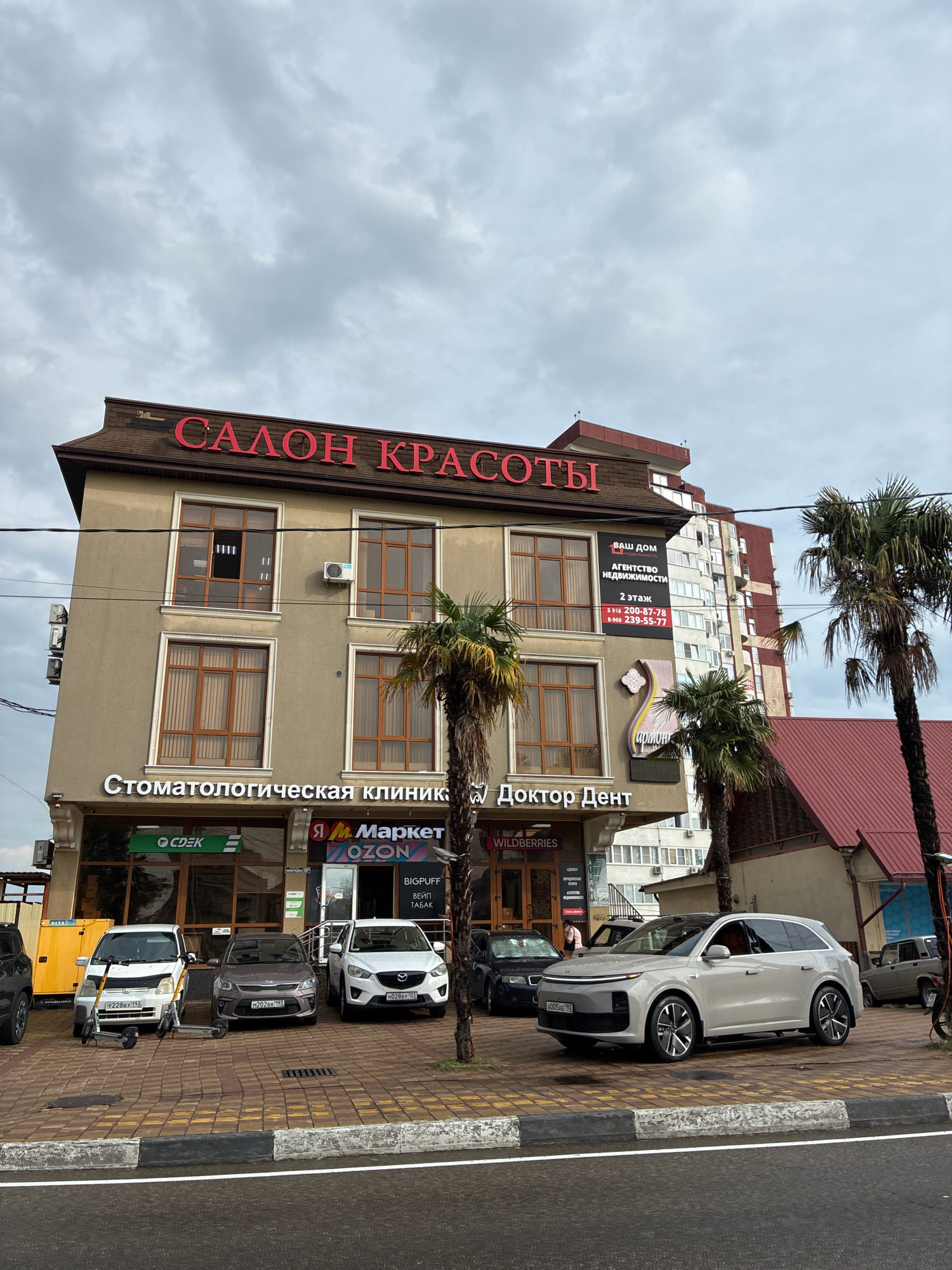 This is a street-level, eye-level, full shot of a commercial building in Sochi, Russia, with a cloudy sky. The building's facade is a light beige color, and it has multiple floors with large, brown-framed windows. Red lettering on the top of the building reads "САЛОН КРАСОТЫ" (Salon of Beauty).
Below the "Salon of Beauty" sign, there are businesses advertised on the facade. A sign in white lettering reads "Стоматологическая клиника" (Dental Clinic) and "Доктор Дент" (Doctor Dent). To the left of the dental clinic, there is a green sign for "CDEK," a logistics company. To the right of the dental clinic, there are signs for "Я Маркет" (Ya Market) and "OZON," both online marketplaces. A smaller sign reads "BIGPUFF ВЕЙП ТАБАК" (BIGPUFF VAPE TOBACCO). There is also an advertisement for "ВАШ ДОМ" (Your Home) real estate agency.
In front of the building, parked cars are visible: a white Kia CX-5, a gray Kia Rio, and a white electric scooter. Further to the right, a modern white SUV is parked. Palm trees are scattered around the scene, some with sparse foliage. A tall, red-bricked apartment building is visible in the background behind the commercial structure. The street in front of the businesses is paved with cobblestones.