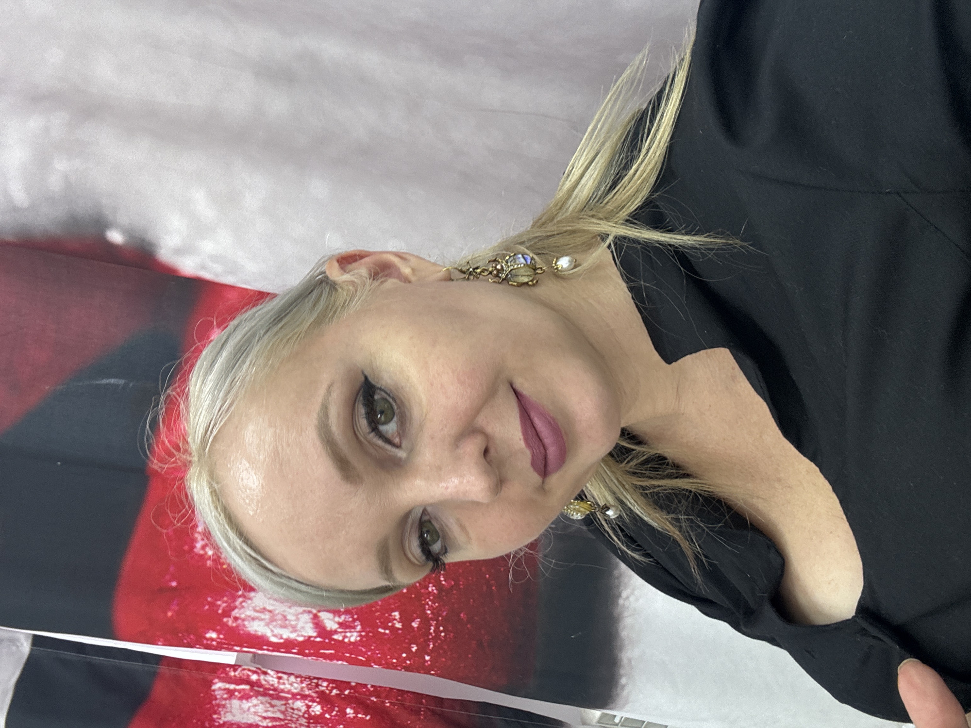 A medium shot shows a woman with blonde hair and makeup. She is wearing a black shirt and ornate earrings. The background is a blurred fabric with red and white patterns. The location is unknown, but the context suggests it is a personal photograph, likely taken indoors.