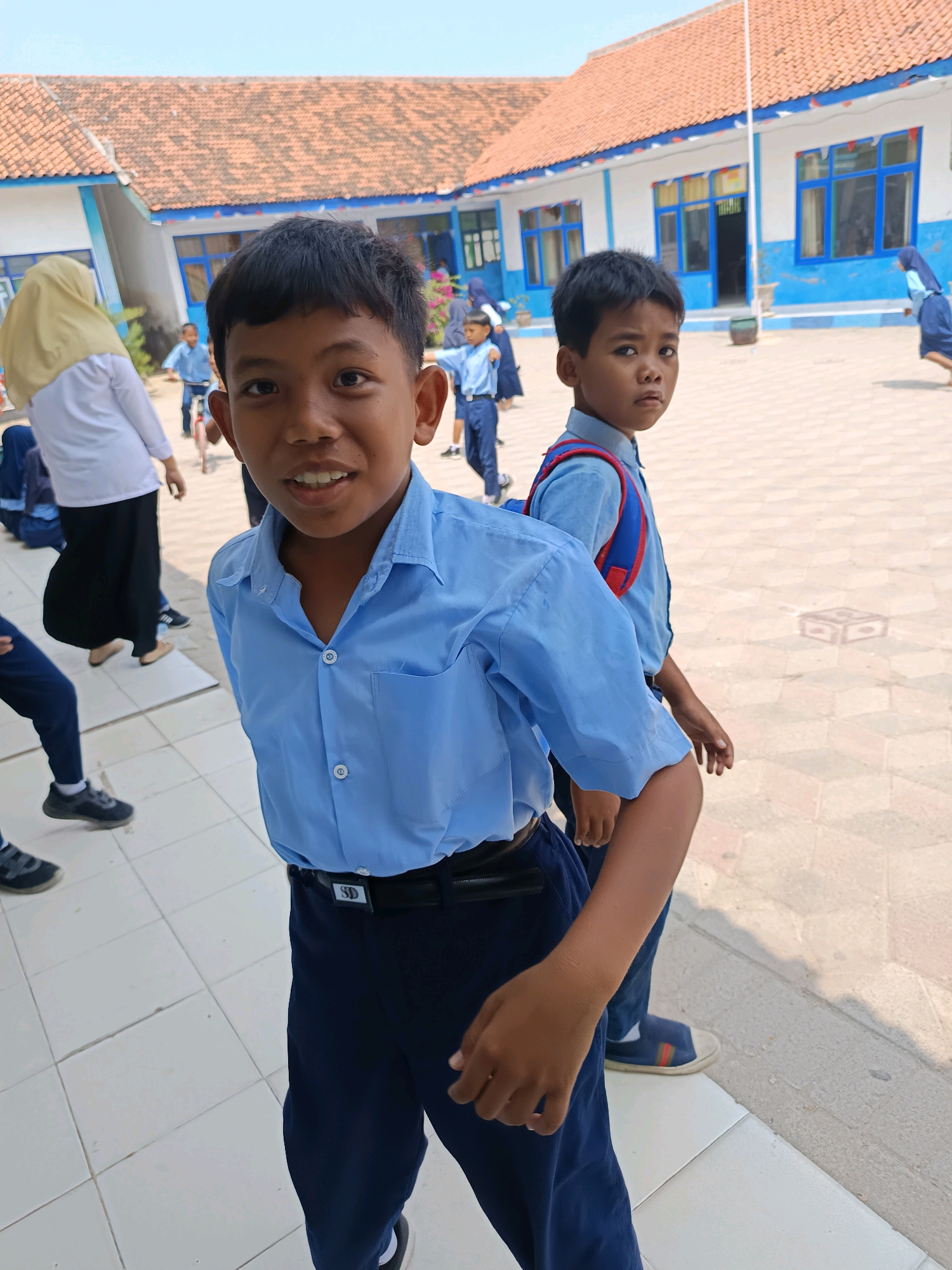 In the city of Labuhan, Indonesia, on a bright, sunny day, two young boys in school uniforms stand in what appears to be a schoolyard. The boy in the foreground, with dark hair and a friendly smile, is wearing a light blue collared shirt tucked into dark blue pants and a black belt with a silver buckle. He is looking directly at the camera. Behind him and slightly to the right, another boy, also in a light blue shirt and dark blue pants, is wearing a red and blue backpack. He is looking to his left. In the background, other children and adults are visible. One adult, wearing a yellow headscarf and a white top with a long black skirt, is standing and observing. The school buildings have white walls with blue trim around the windows, and the roof is made of red tiles. The ground is paved with light-colored square tiles. The overall scene depicts a typical day at an Indonesian school.
