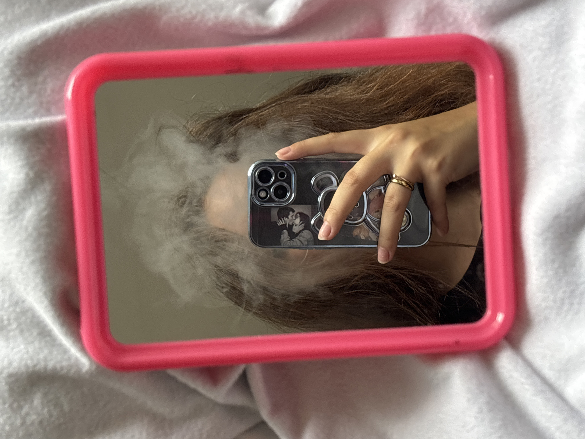 The media file shows a close-up reflection in a pink-framed mirror. The reflection reveals a person holding a smartphone, which is being used to take the picture. The person's hand, adorned with two gold rings, is visible, along with strands of brown hair. A significant amount of white smoke or vapor is also visible, obscuring part of the person's face and head in the reflection. The smartphone case has a decorative design featuring a stylized bear or character. The mirror is placed on a white, textured surface, likely a bed or blanket, which is draped and folded. The lighting suggests an indoor setting, possibly daytime due to the brightness. There is no visible text in the image. The context provided indicates the location is Göttingen, Germany.