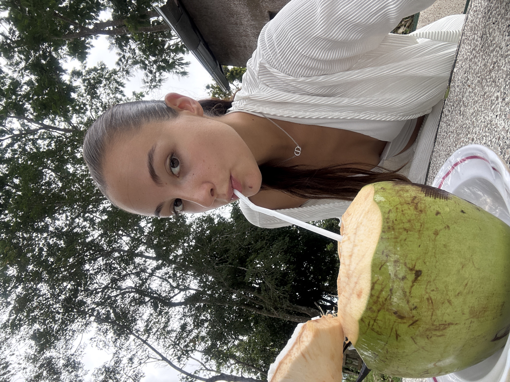A young woman is drinking from a coconut with a straw. She is wearing a white top and a silver necklace with a "D" pendant. The background shows trees and possibly a building overhang. There's a table with a white plate and the coconut on it. The location is identified as Karon, Thailand, based on geographical and environmental details consistent with the region.
